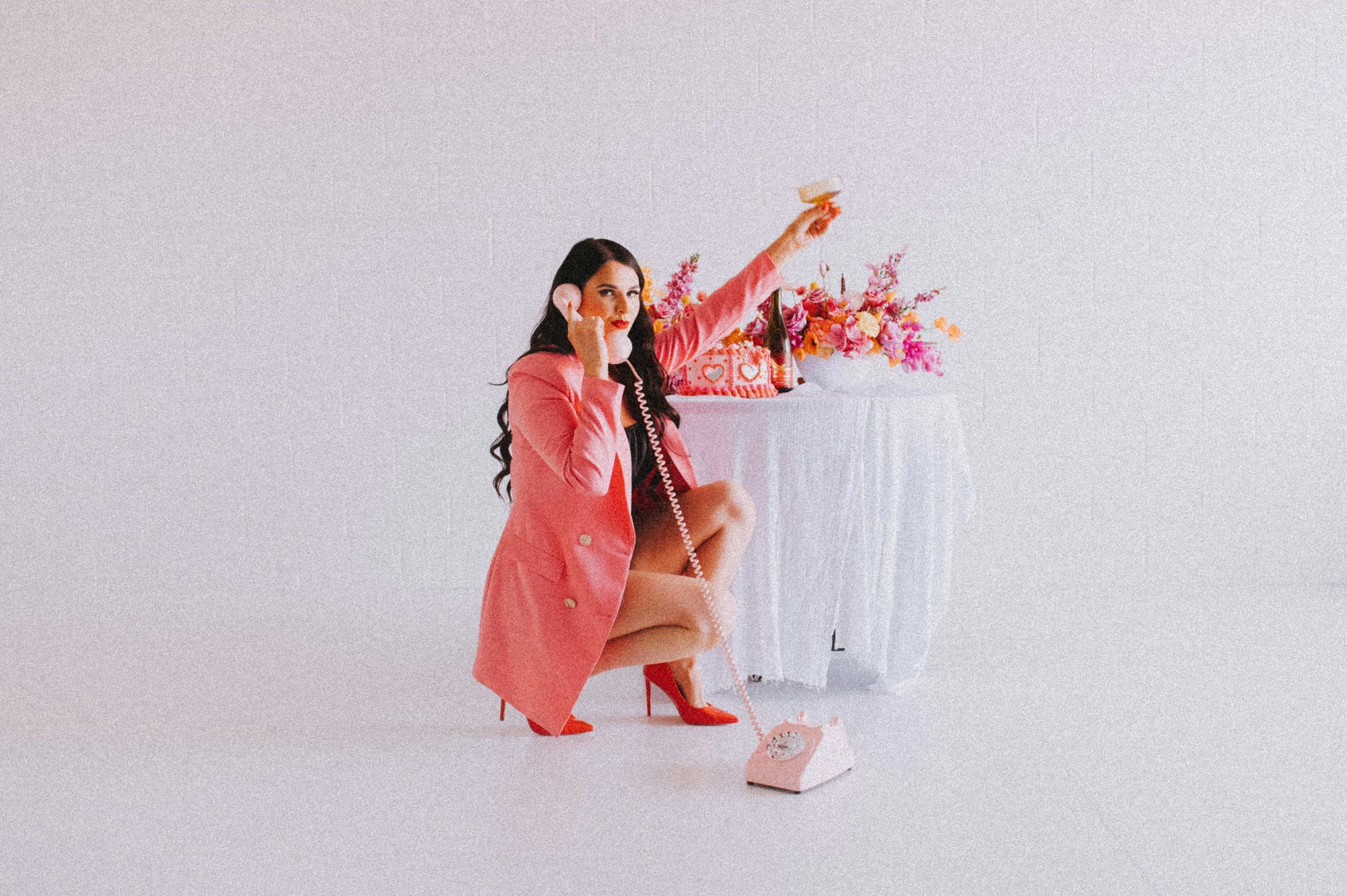 A woman in a pink coat and red high heels squats on a white floor, holding a vintage pink telephone with the cord stretched towards the floor. She is posing next to a round table draped with a white cloth, decorated with a large bouquet of pink, orange, and yellow flowers and a pink cushion with hearts. She raises a glass in her right hand and touches her face with her left hand, with a neutral expression and long dark hair.