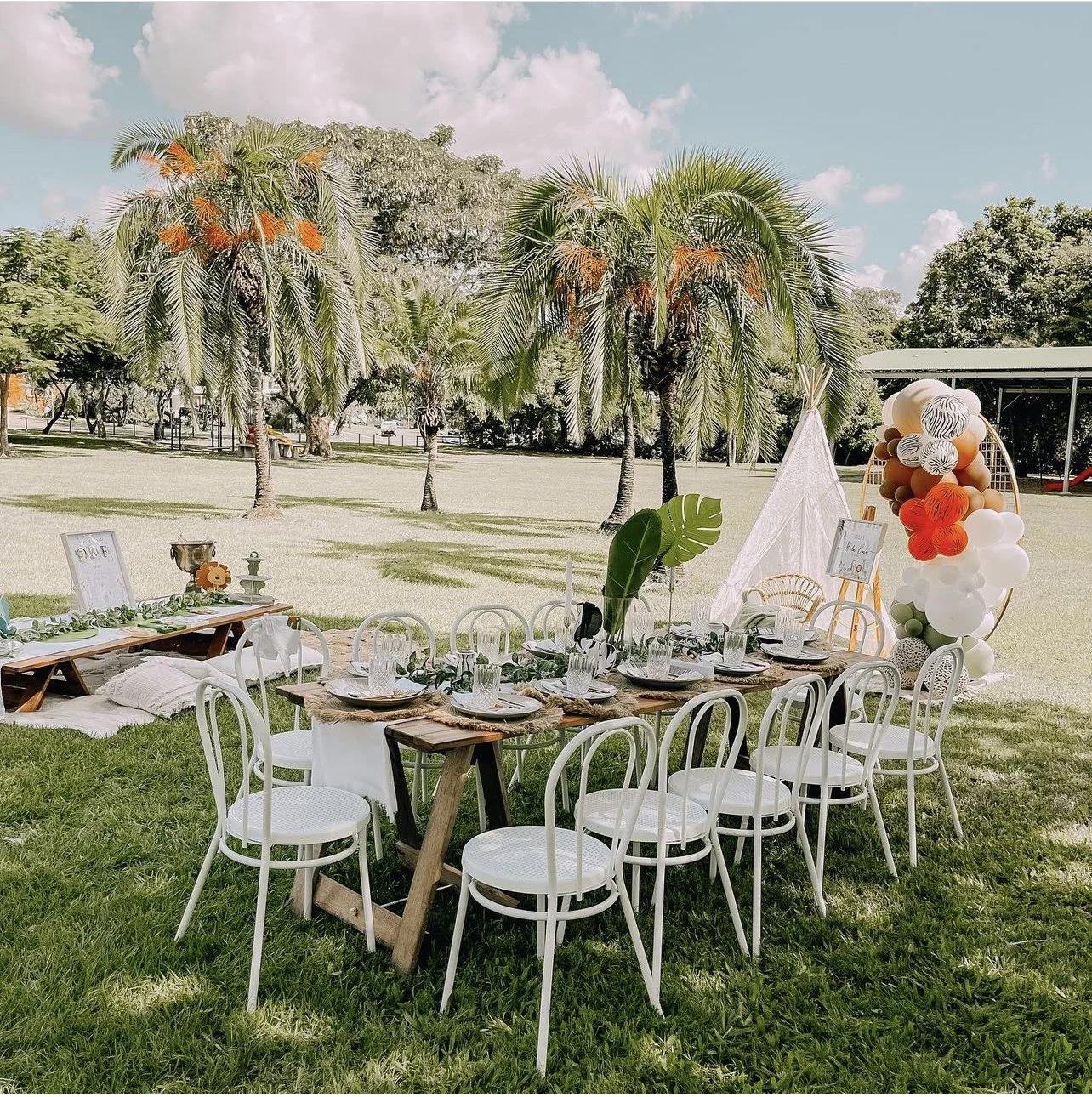Outdoor party setup with a long wooden table, white chairs, tropical plants, and colorful balloon decorations under a partly cloudy sky.