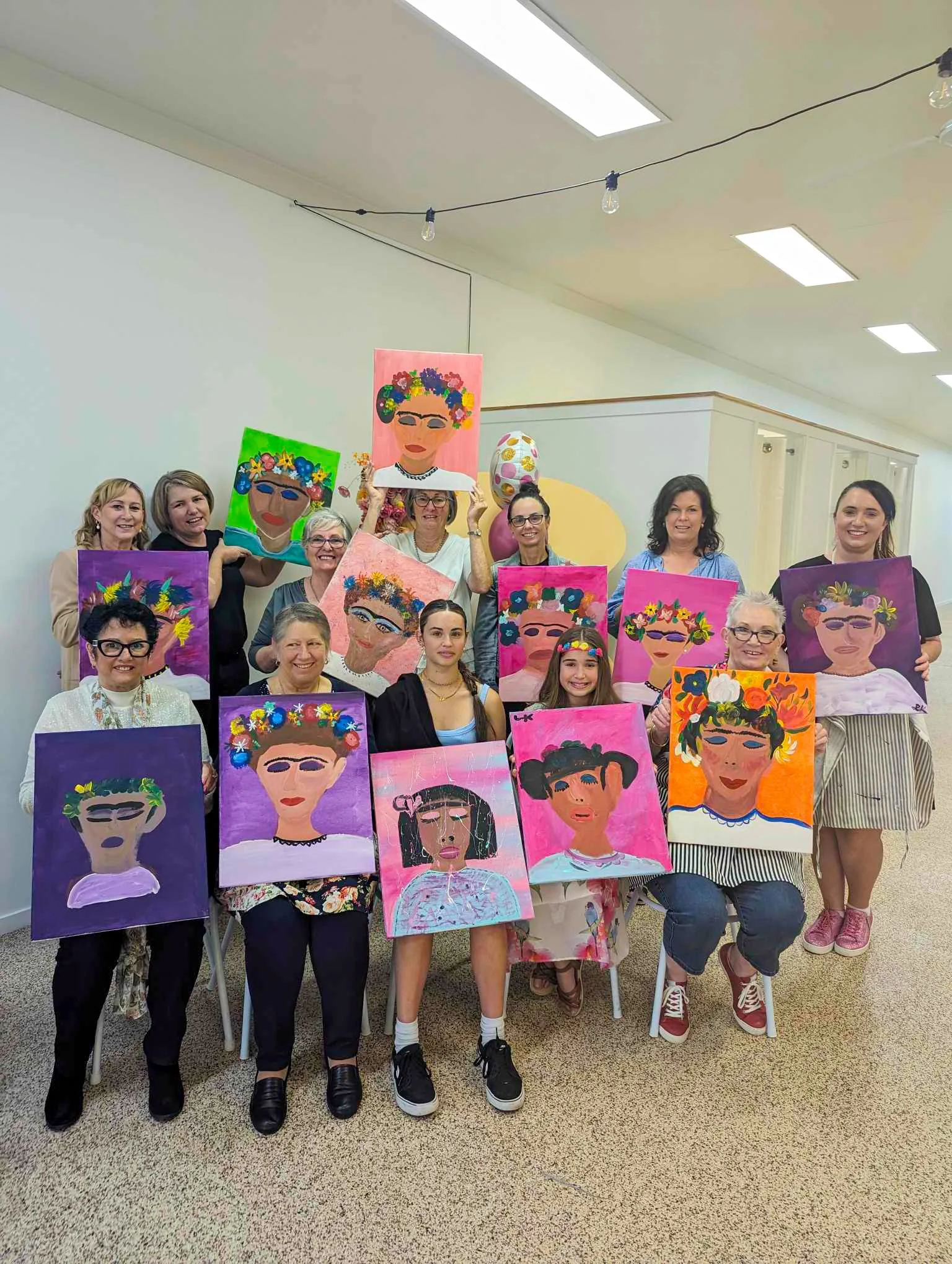 Group of women and girls holding colorful portrait paintings of a woman wearing a floral crown, in an indoor setting with string lights and a ceiling light.