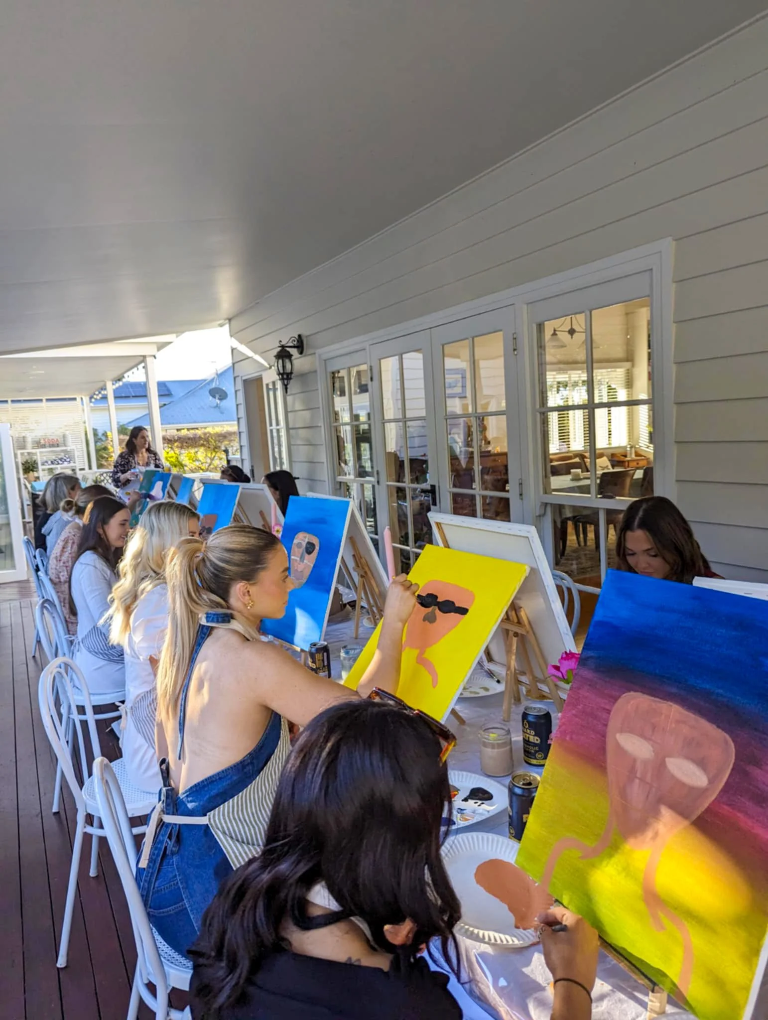 People painting on canvases during an outdoor group painting event on a covered porch.