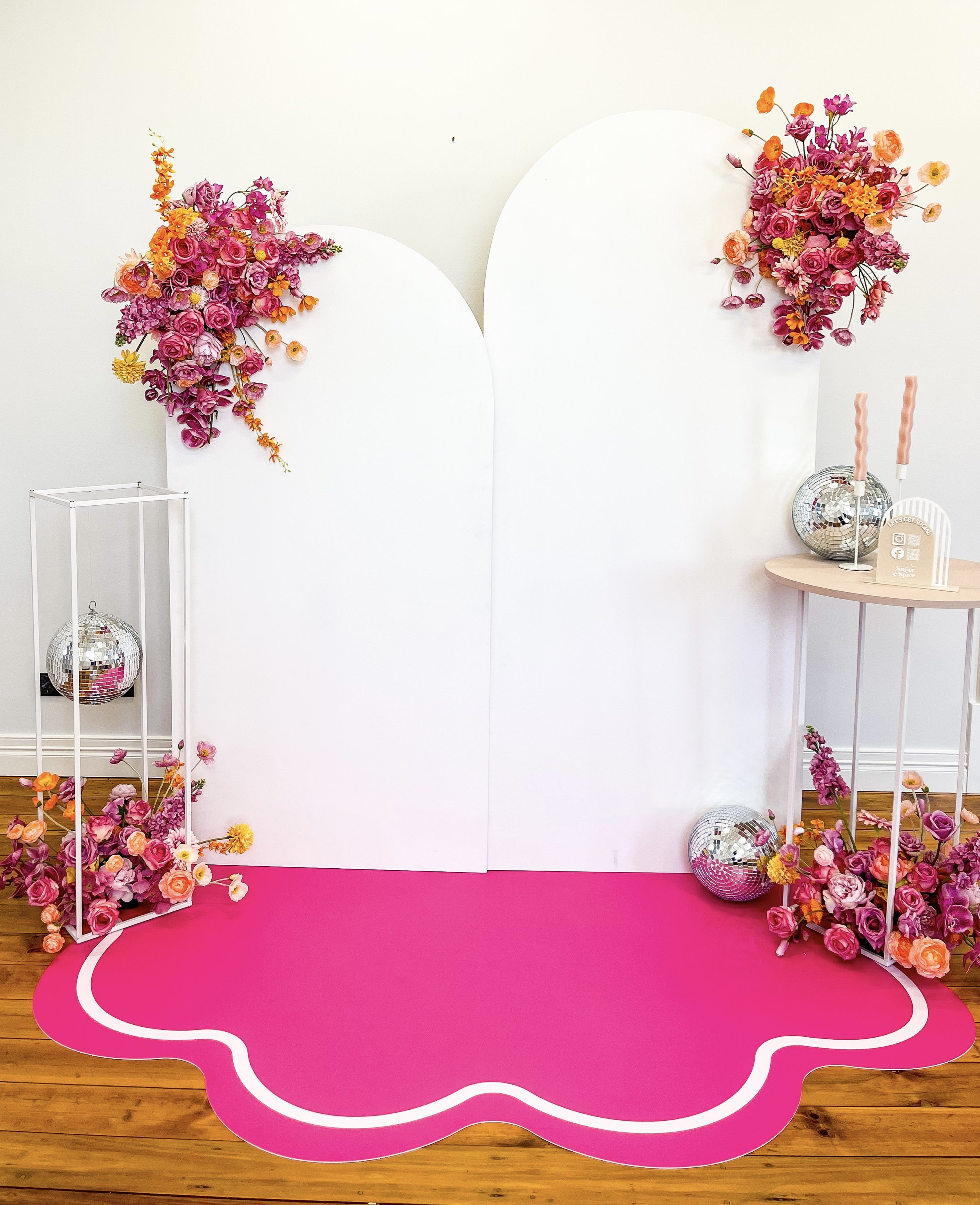 Decorative backdrop with white panels adorned with pink and purple floral arrangements, pink and orange accents, disco balls, and pink carpet on wooden floor.