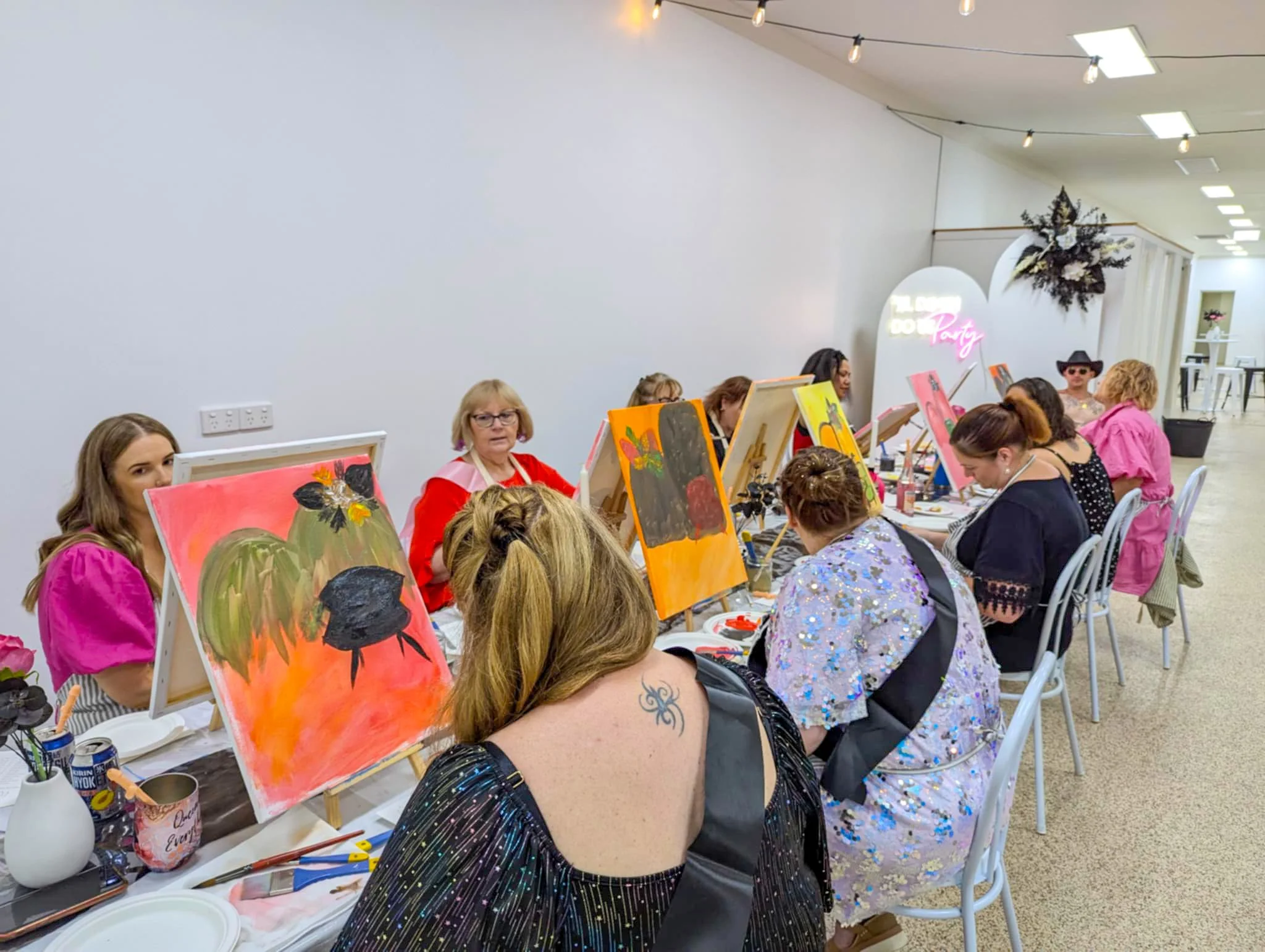 A group of women participating in a painting class, seated at a long table with easels, colorful paintings, and art supplies, in a bright room with string lights and decorative signs.