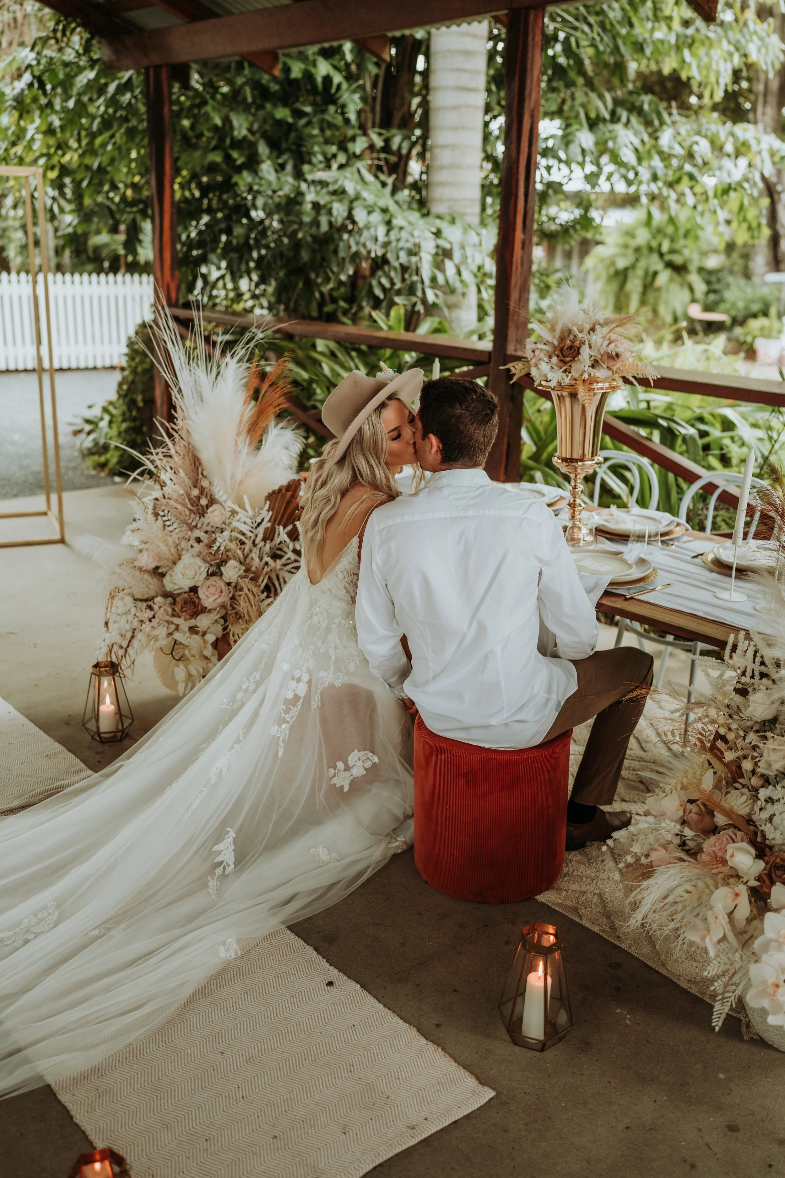 A bride and groom share a kiss at their wedding reception, seated at a decorated table with floral arrangements and candles, in an outdoor garden setting.