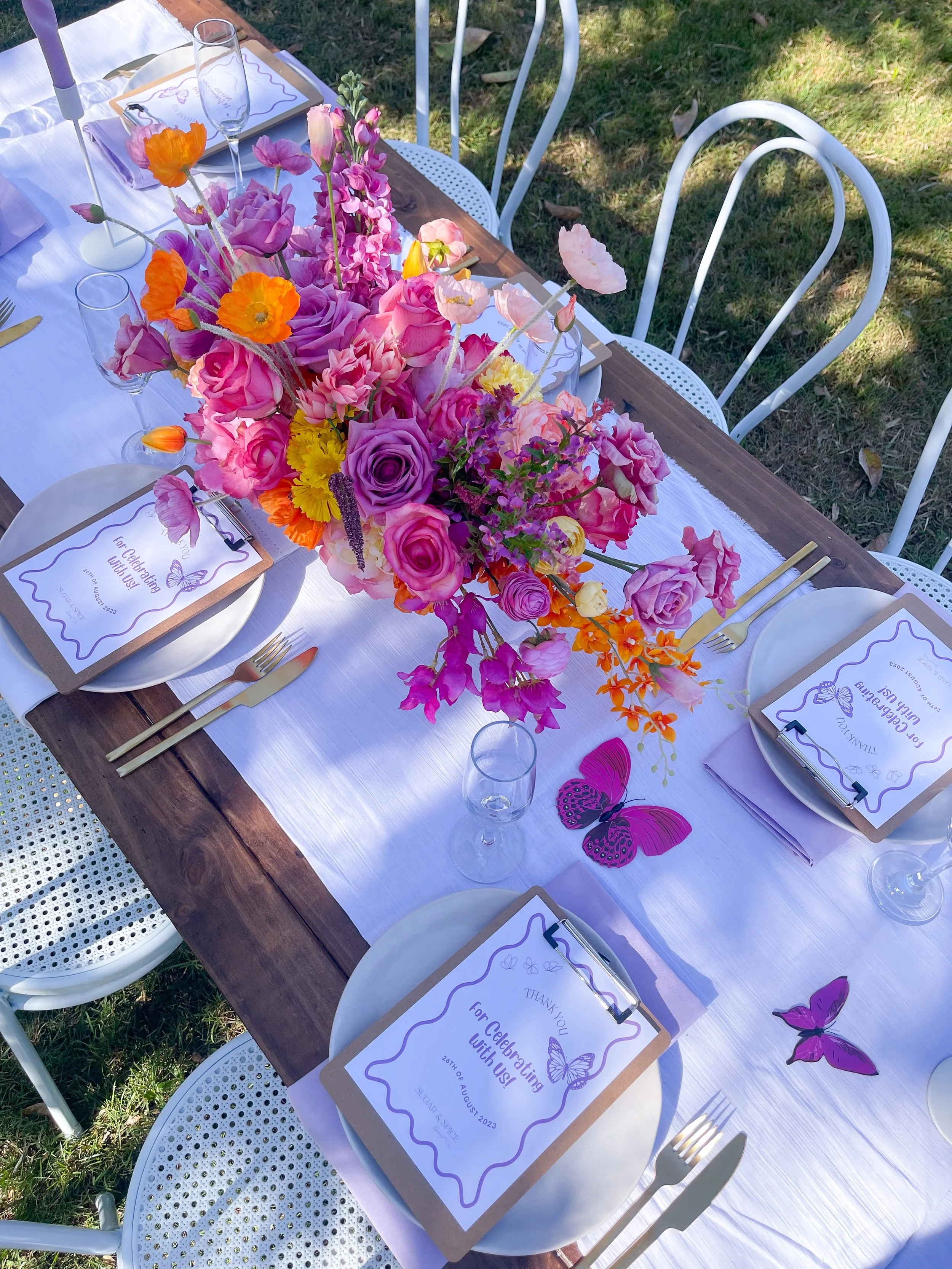 A table set for an outdoor celebration with a white tablecloth, a brown wooden centerpiece with pink, purple, orange, and yellow flowers, and butterfly decorations. Each place setting has a white plate, gold utensils, a champagne flute, and a card th