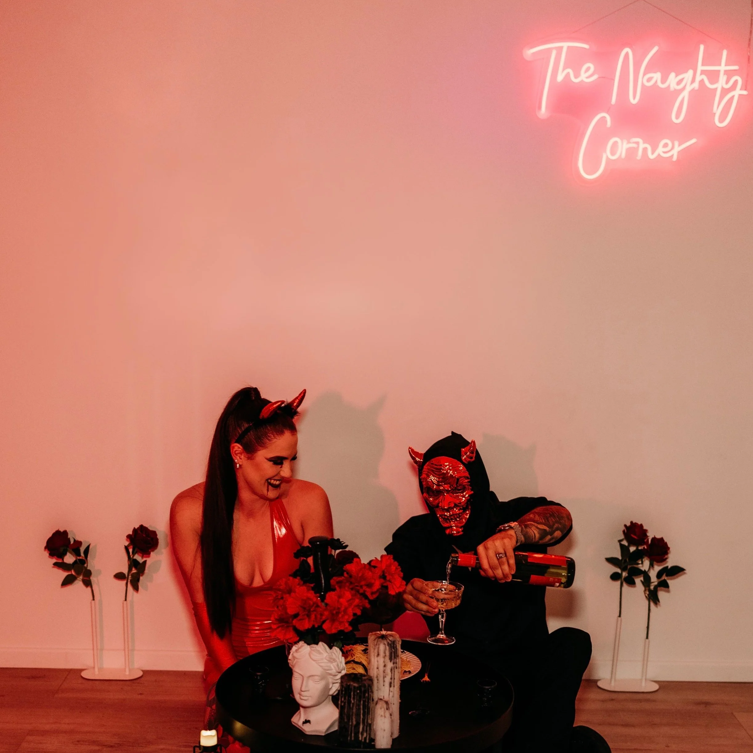 Two people dressed in devil costumes sitting at a table with flowers, candles, and decor. One person is wearing a red outfit with devil horns and has a long dark ponytail. The other has a red devil mask and black hoodie, pouring a drink. Pink neon sign reading 'The Naughty Corner' is on the wall behind them.