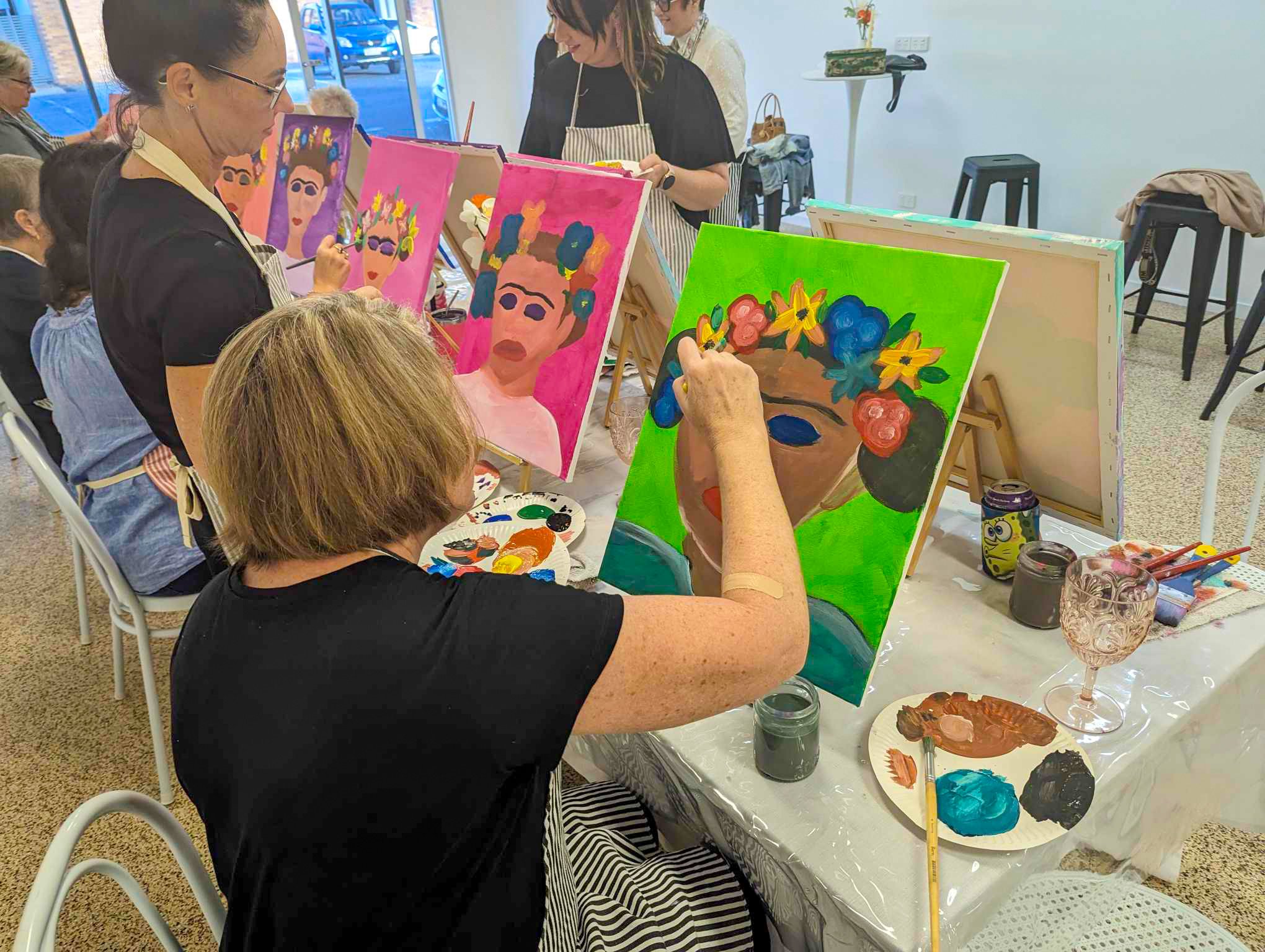 People participating in an art class, painting colorful portrait-style images of women on canvas, with painting supplies and others observing.