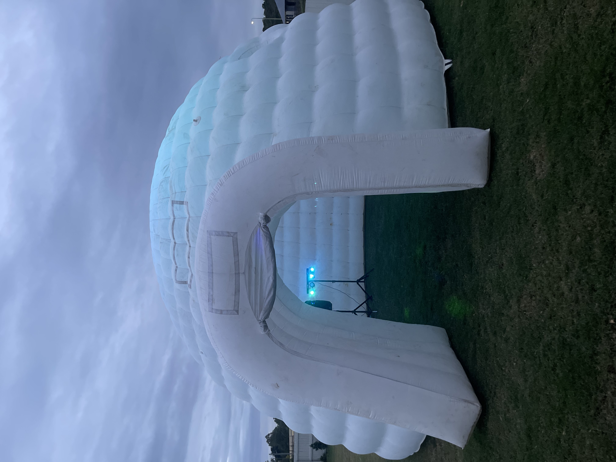 A white inflatable structure resembling a snow cave or igloo is set up on a grassy area during dusk, with a small electronic device emitting blue and green light nearby.