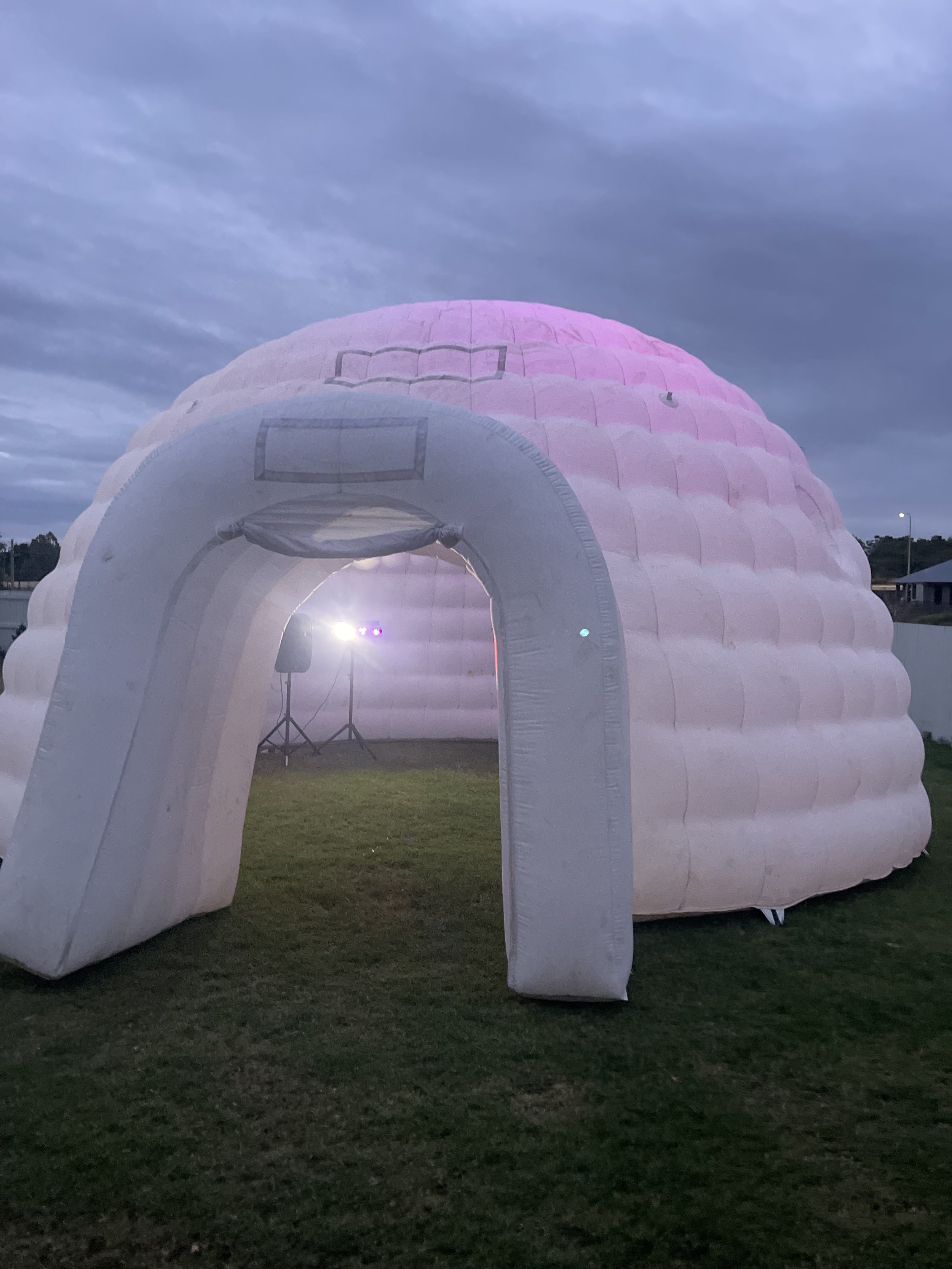 An inflatable igloo-shaped structure with a pink and white color design, set up outdoors on a grassy area during the evening, with a small stage and lighting equipment visible inside.