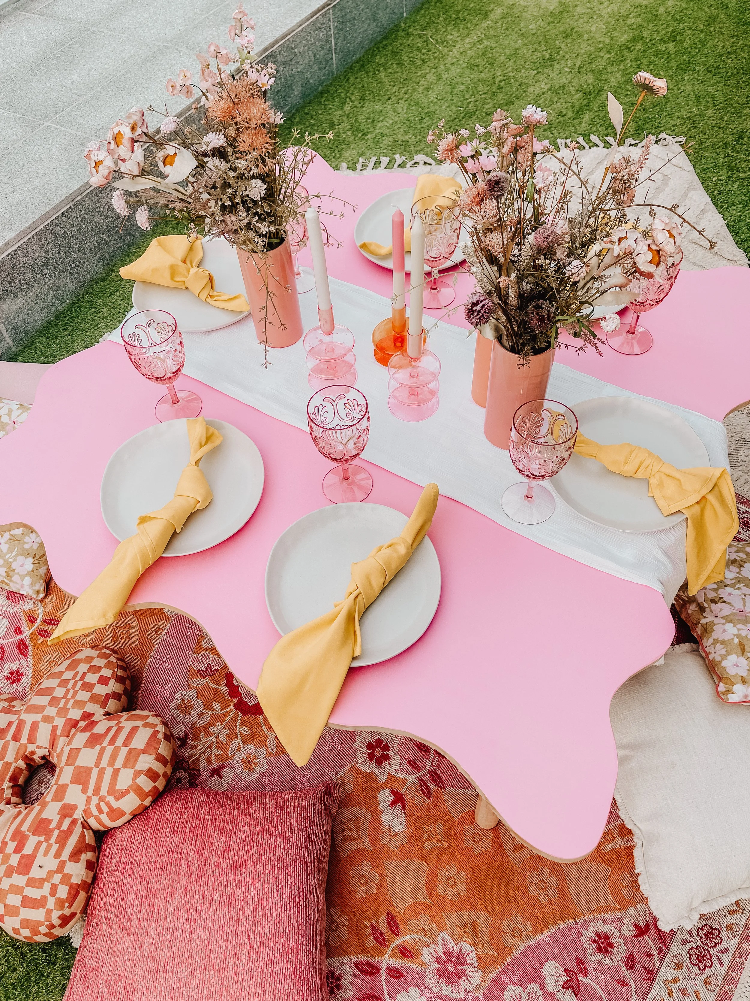 Decorative outdoor dining table set with pink tablecloth and floral centerpiece, featuring white plates with yellow napkins, pink glassware, and candles, surrounded by pillows and rugs on a grassy area.