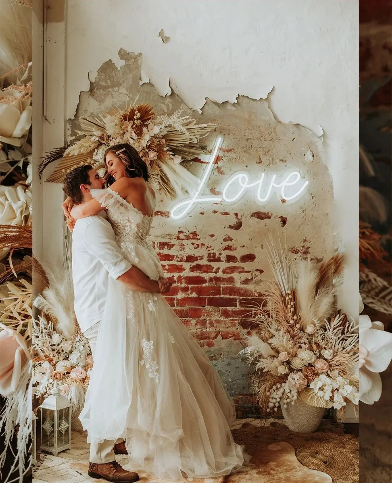 A couple in wedding attire, with the groom lifting the bride, standing in front of a rustic brick and plaster wall with a neon 'Love' sign and floral arrangements.