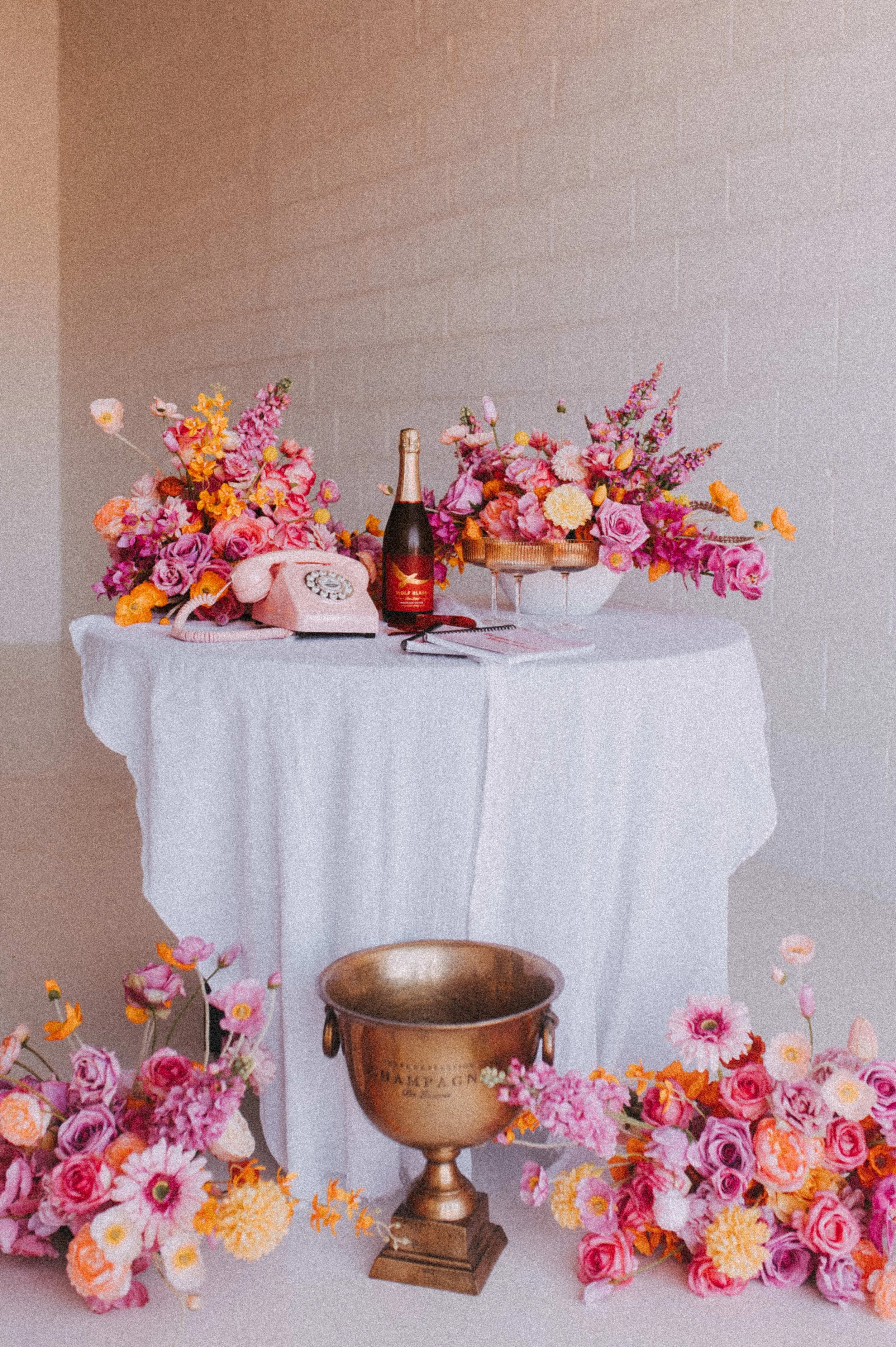A white-tabled display with pink and orange flowers, a pink rotary phone, a bottle of champagne, and a small notebook. There are additional pink and orange flowers on the floor and a gold trophy cup in front of the table.
