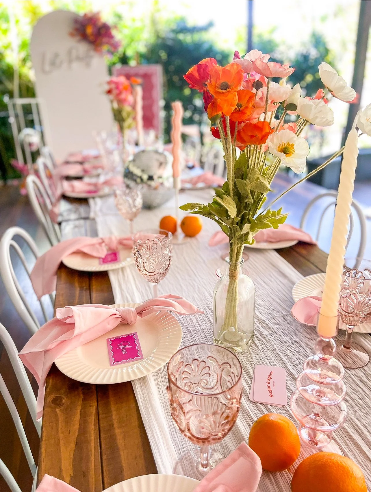 A decorated table set for a celebration with pink-themed tableware, pink napkins, vintage pink glasses, a floral centerpiece with pink, orange, and white flowers in a glass vase, and a pink card labeled 'Sugar & Spice'.