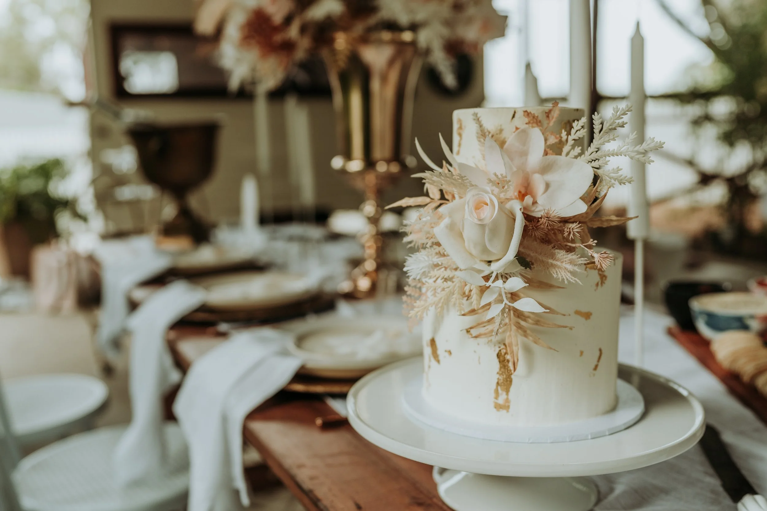 A white wedding cake decorated with white flowers and foliage, placed on a cake stand on a dining table set with plates, napkins and candles, in a well-lit room with windows.