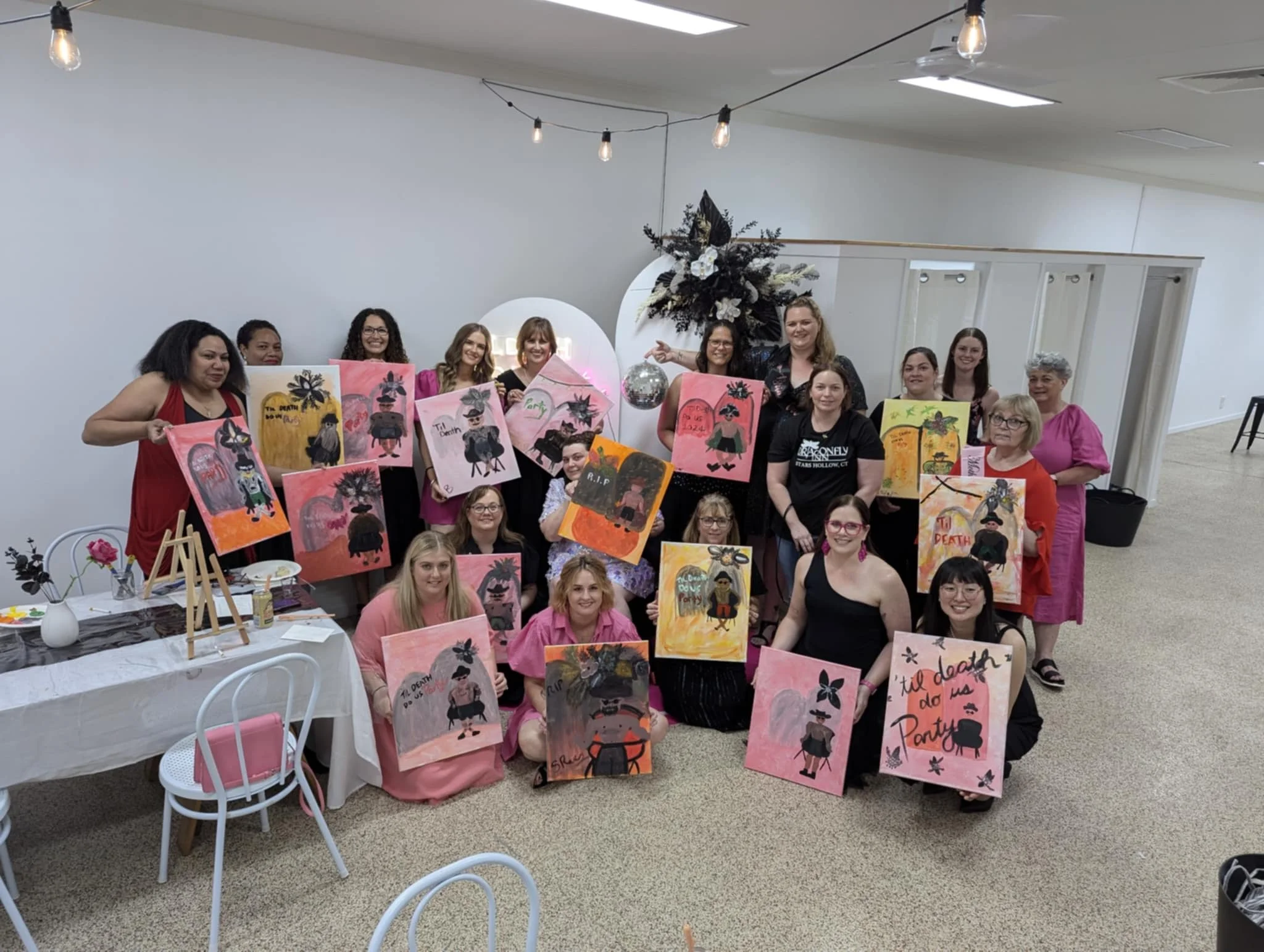 Group of women holding paintings at an art event in a decorated indoor space.