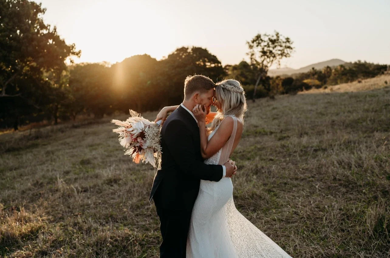 A bride and groom in a field at sunset, embracing and touching foreheads, with the bride holding a bouquet of flowers.