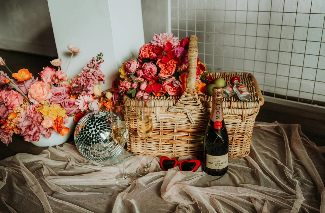 A display of flowers, champagne, and decorative items on a textured fabric surface, including a large floral arrangement, a bottle of Moët & Chandon champagne, two glasses of champagne, a disco ball, and red sunglasses.