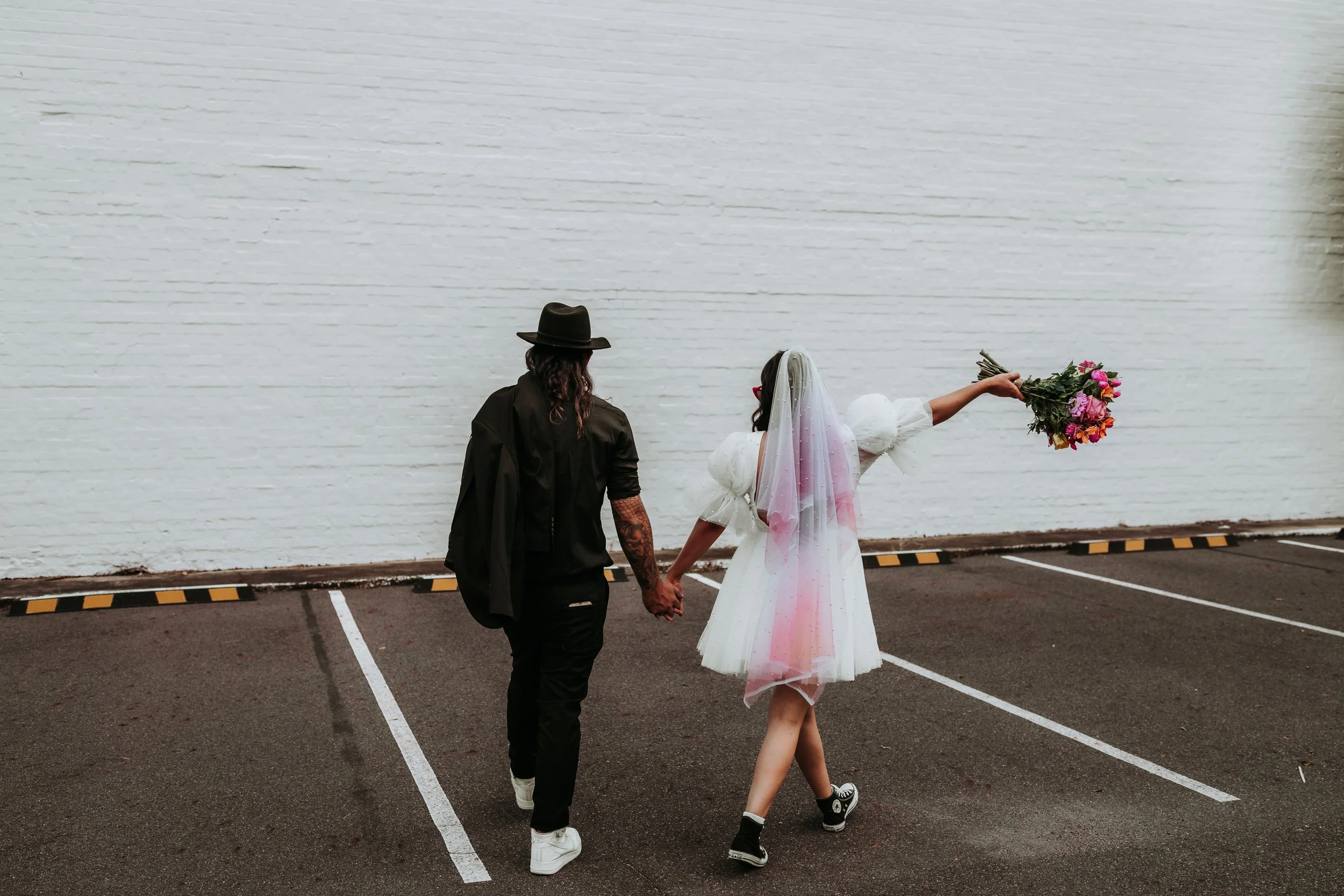 A couple, one in casual black attire and the other in a white wedding dress with a veil, holding hands in an empty parking lot. The woman in the wedding dress is holding a bouquet of colorful flowers and extending her arm with the bouquet.