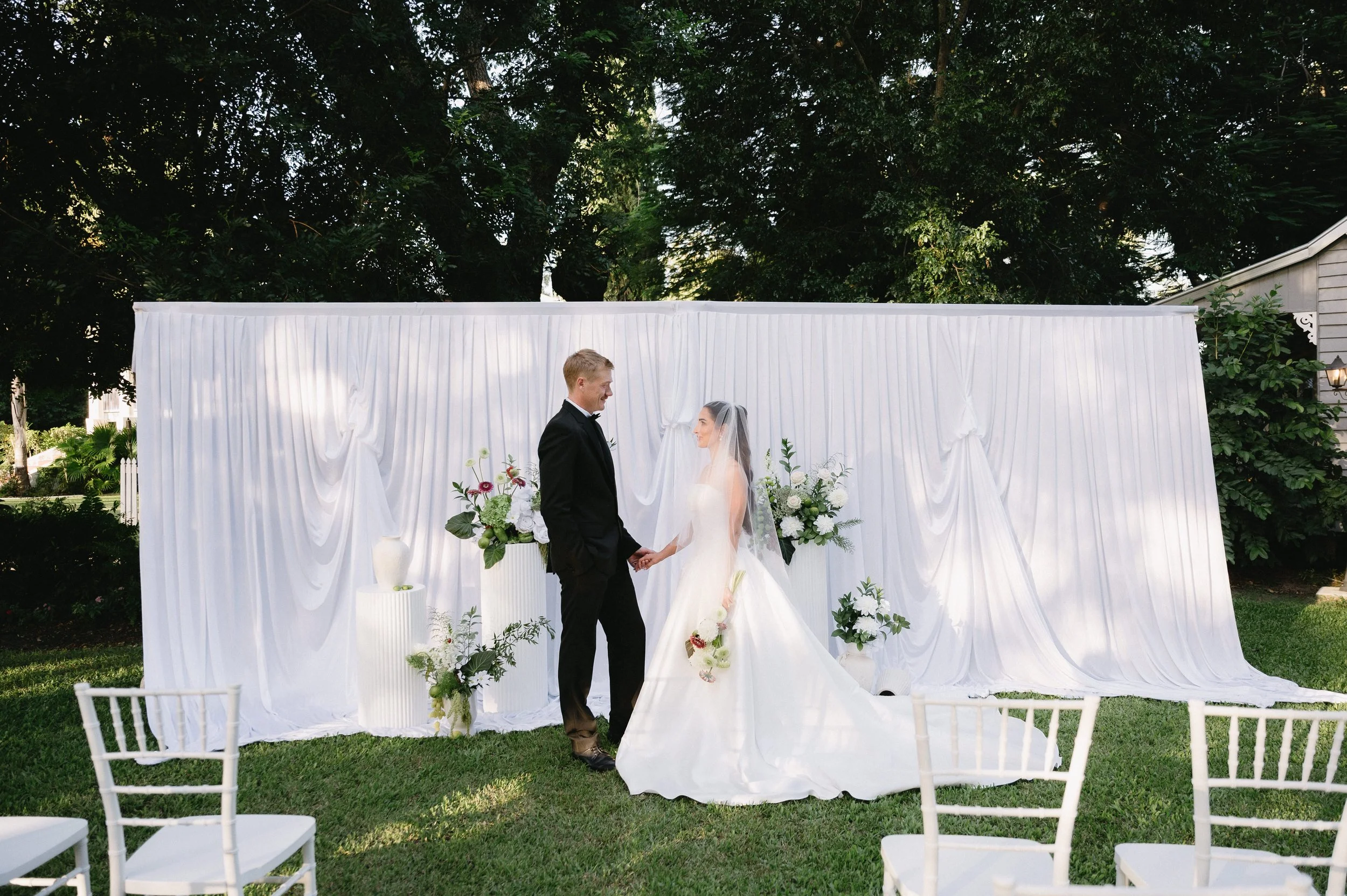 Bride and groom holding hands during their wedding ceremony outdoors, with a white draped backdrop and flowers