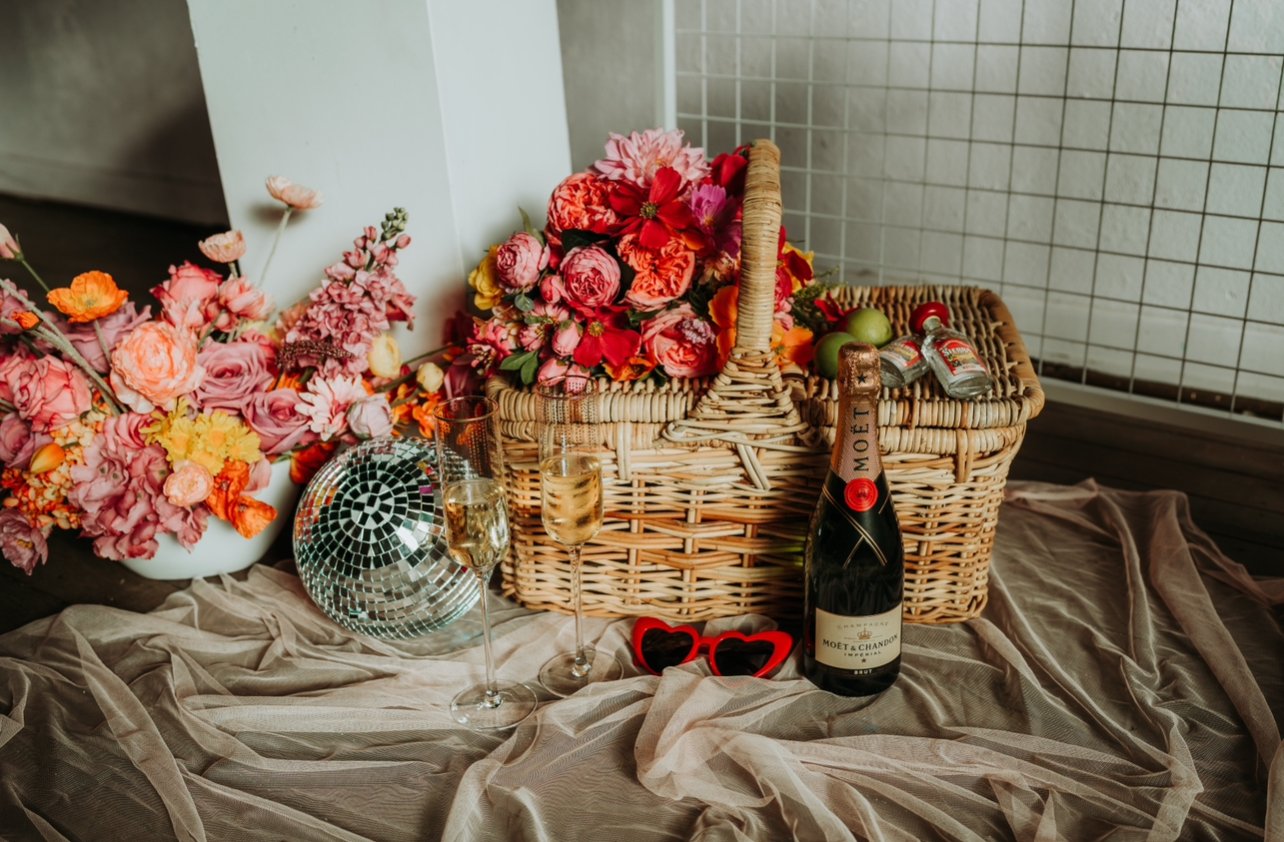 A bouquet of pink and red flowers with a wicker picnic basket, a bottle of Moët & Chandon champagne, two champagne glasses, a disco ball, and red sunglasses on a draped beige fabric.