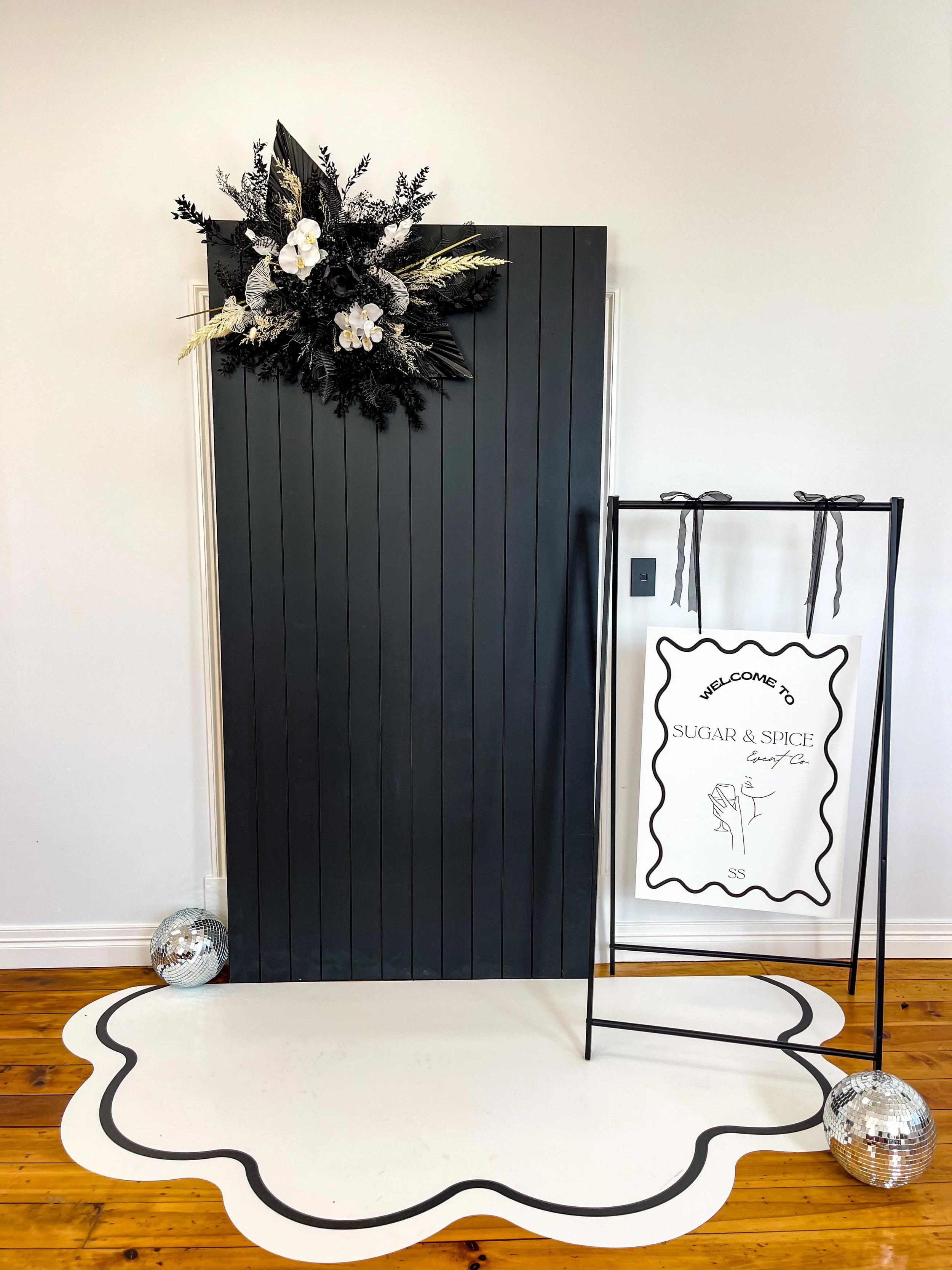 Decorative black wooden door with a floral arrangement of black, white, and gold foliage and orchids on top. A white and black scalloped rug is on the wooden floor with two disco balls nearby. To the right, a black metal frame holds a white sign with black lettering that reads, "Welcome to Sugar & Spice Event Co." with a stylized illustration of a hand holding a drink.