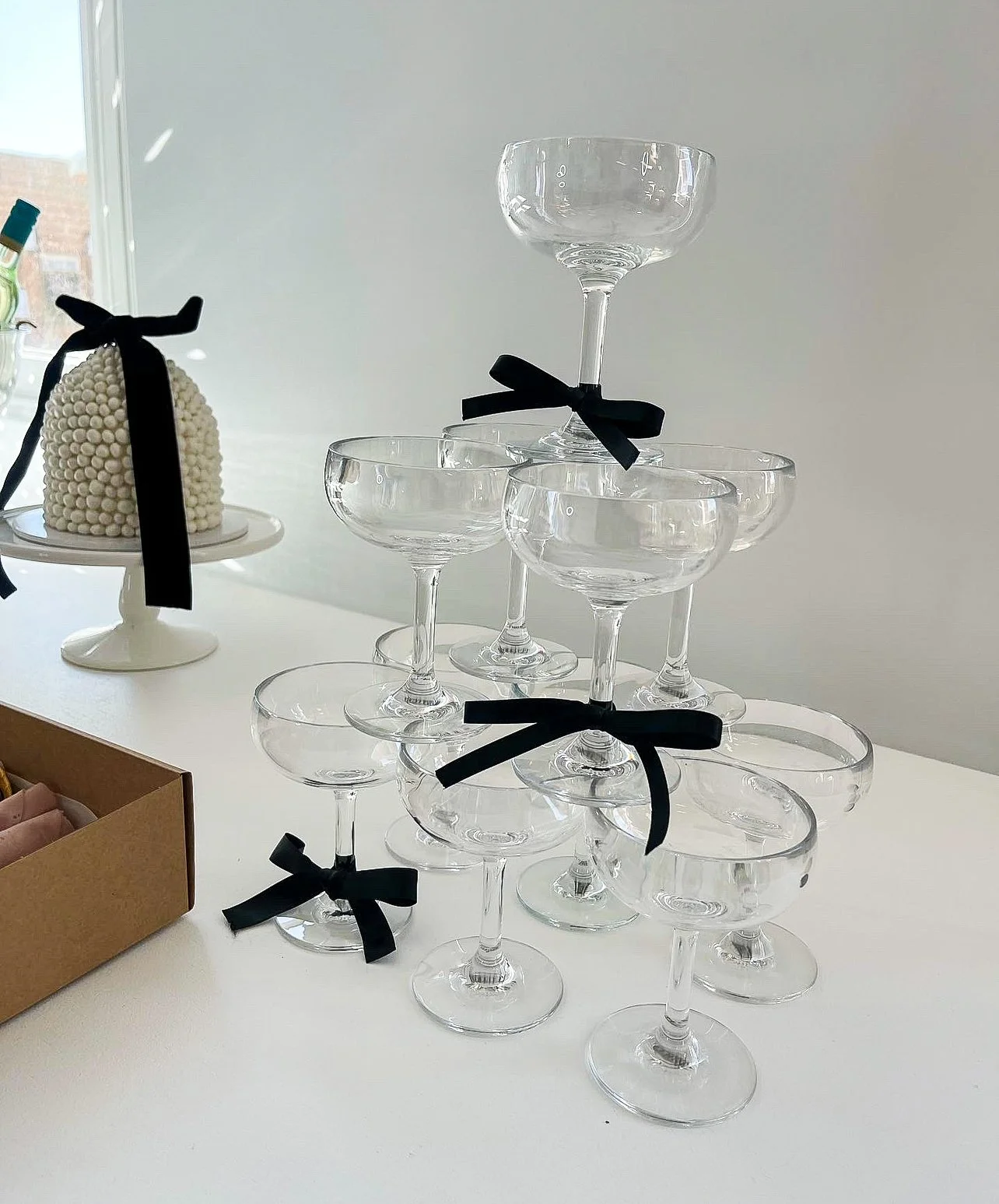 Collection of empty champagne glasses stacked in a pyramid with black ribbons tied around them, placed on a white surface with a white wall in the background.