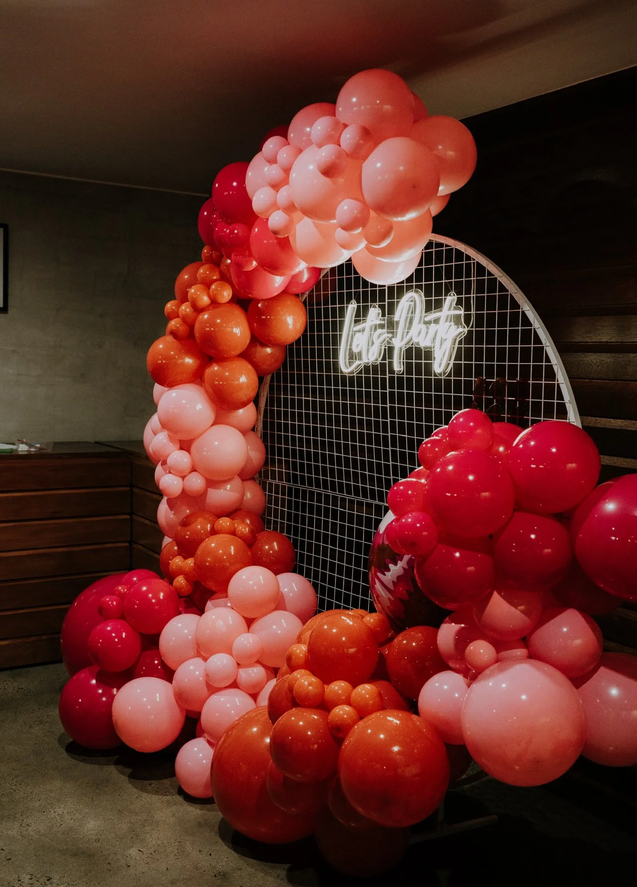 Decorative balloon arch with various sizes in shades of pink, red, and orange, with a neon sign in the background that says 'Let’s Daff' against a metal grid backdrop.