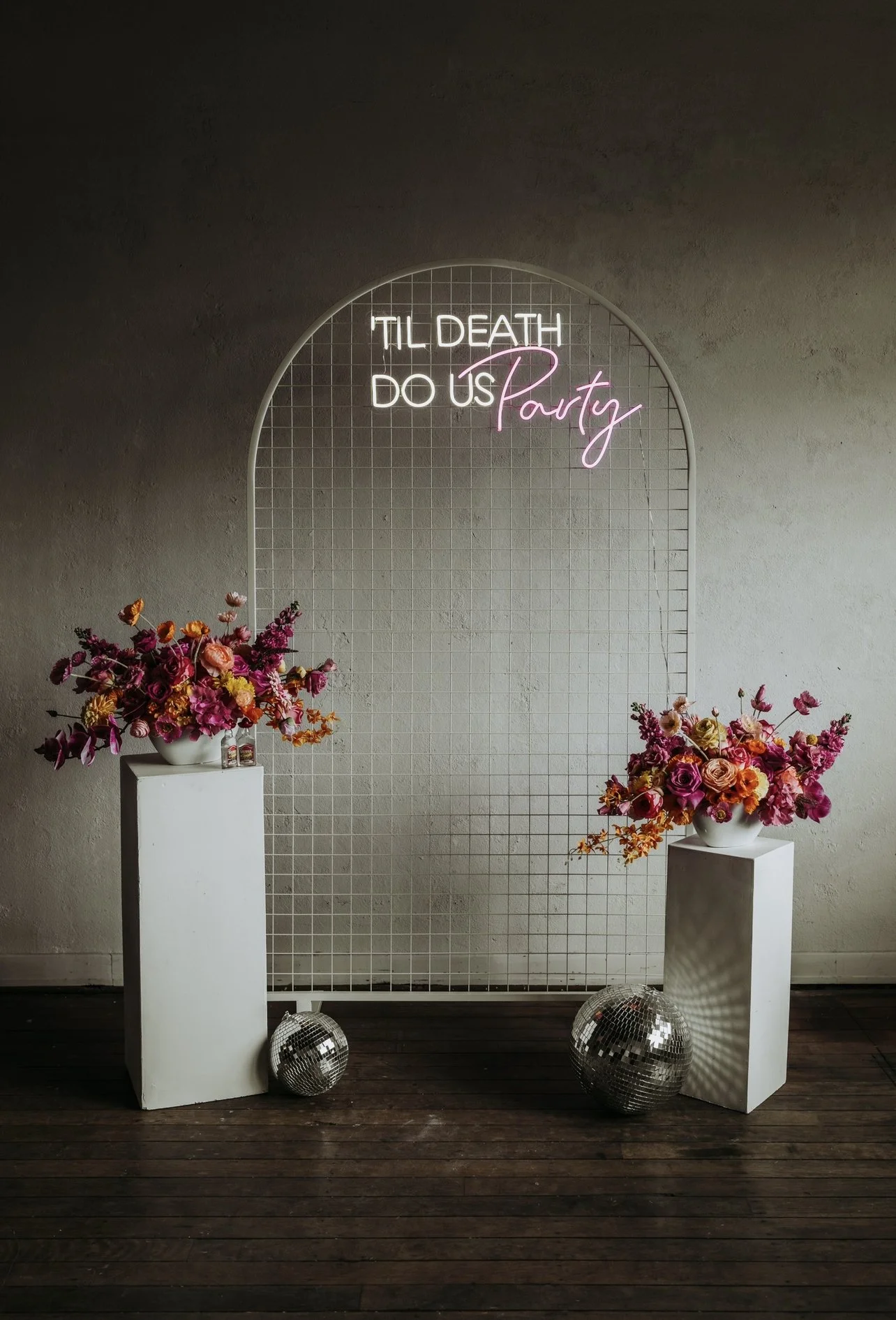 A decorative party setup with two white pedestals holding floral arrangements, disco balls on the floor, and a wire grid backdrop with a neon sign that reads 'Til Death Do Us Party' with 'Party' in pink script.