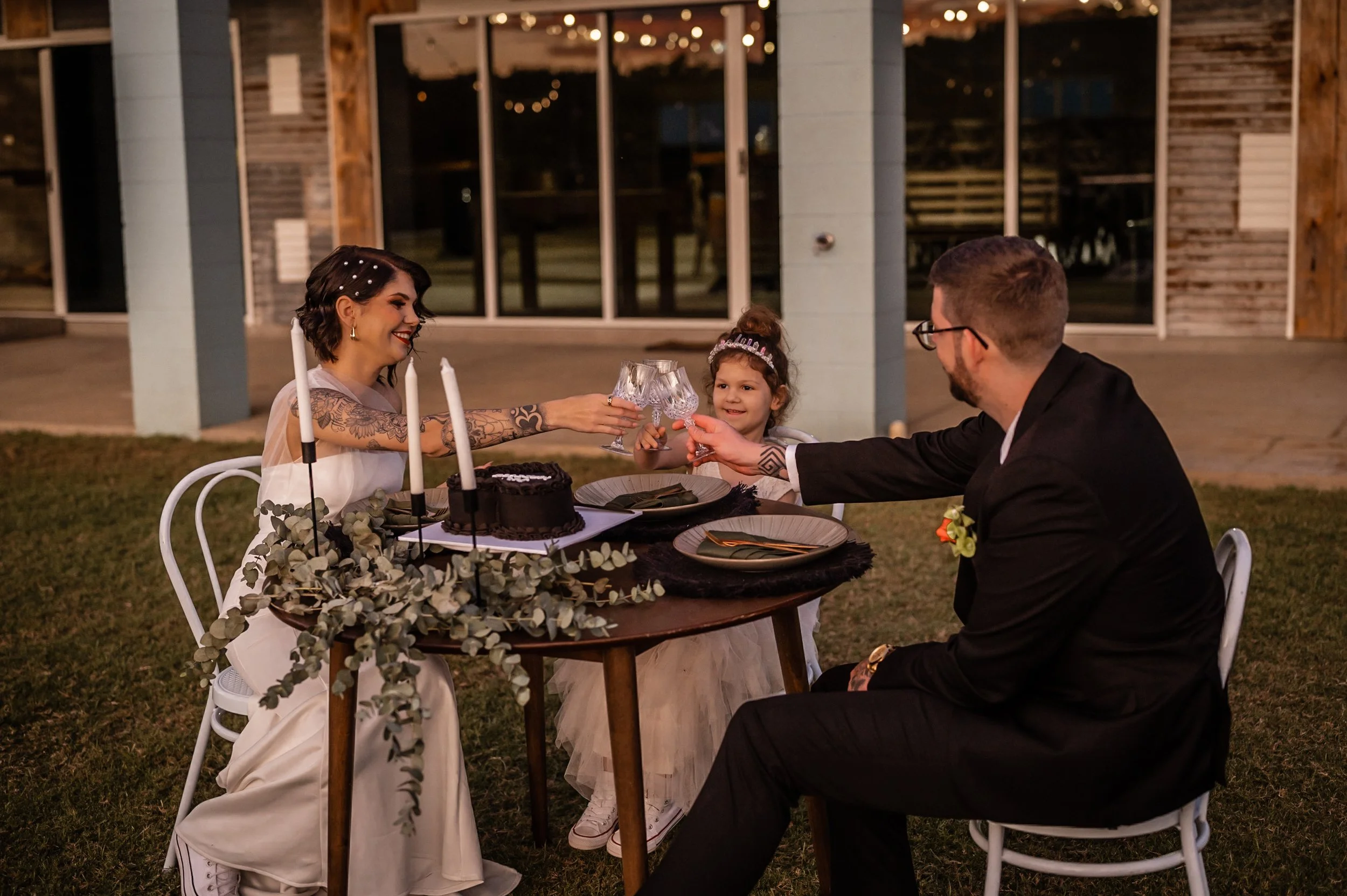 A family celebrating a birthday outdoors at night, with a woman, man, and young girl toasting with wine glasses at a table with a chocolate cake, candles, and decorative greenery.