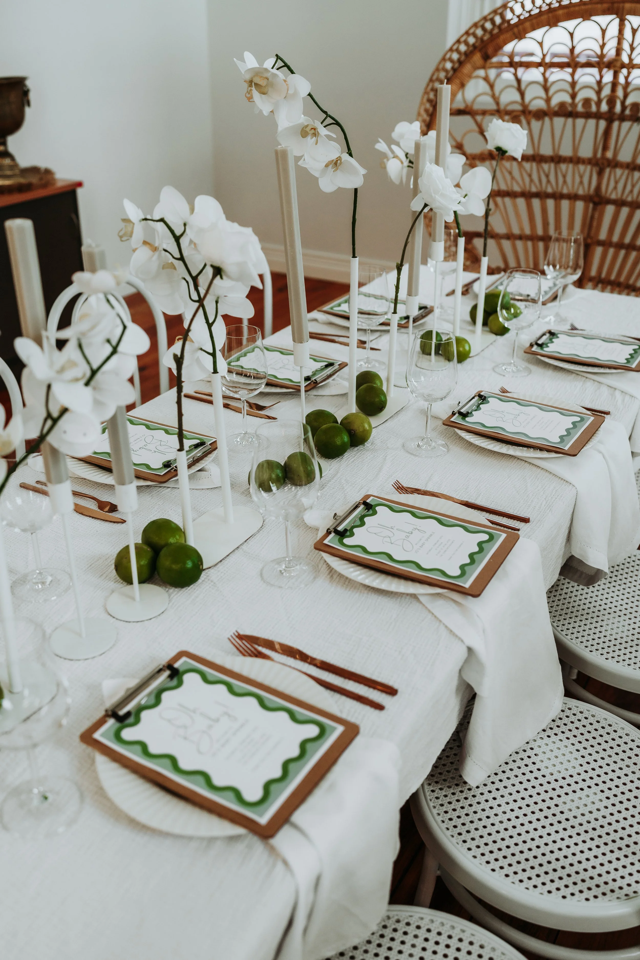 A dining table set for a celebration with white orchids, green apples, candles, and printed menus on white plates, with white chairs surrounding the table.