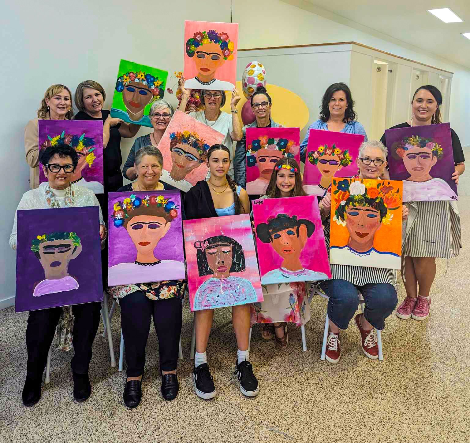 Group of women and a girl holding colorful paintings of Frida Kahlo self-portraits, depicting vibrant backgrounds and floral crowns, in an indoor setting.