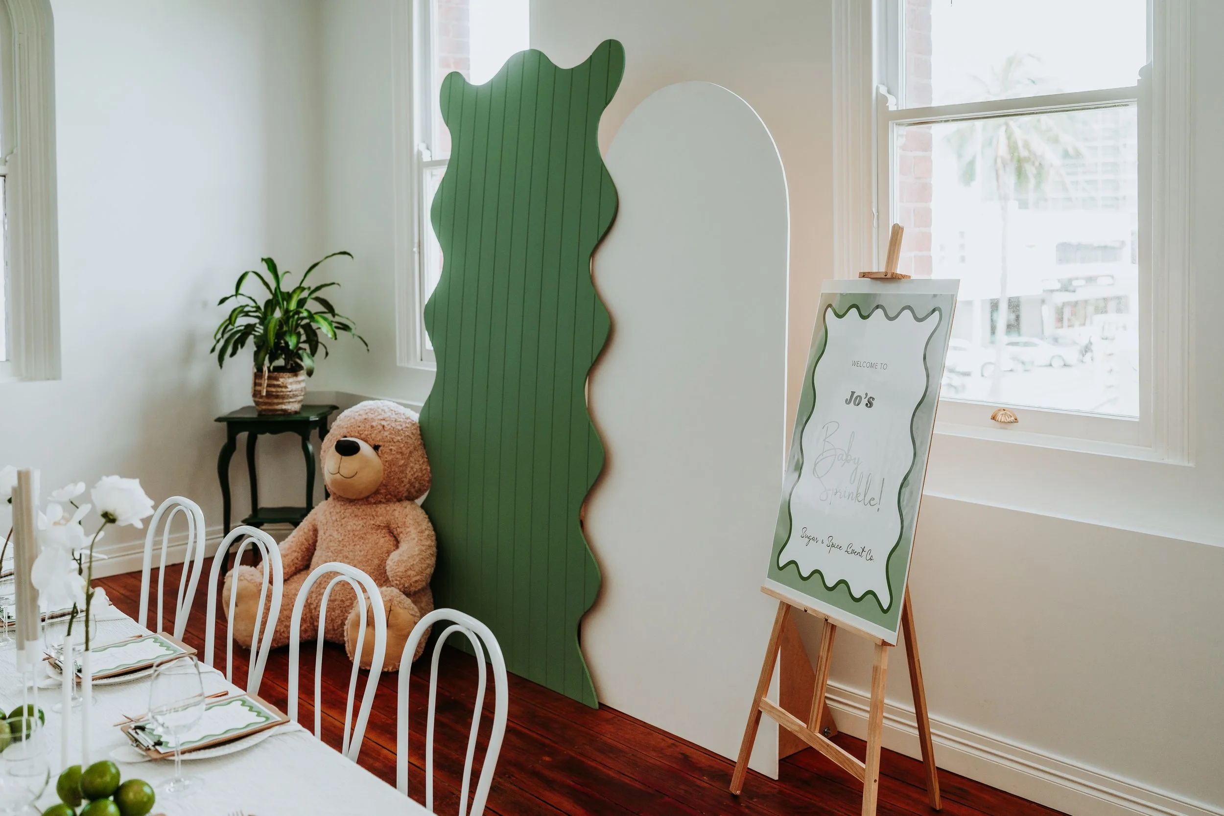 Baby shower setup with a teddy bear, green and white decorations, and a welcome sign that reads, "Jo's Baby Sprinkle!" near a window