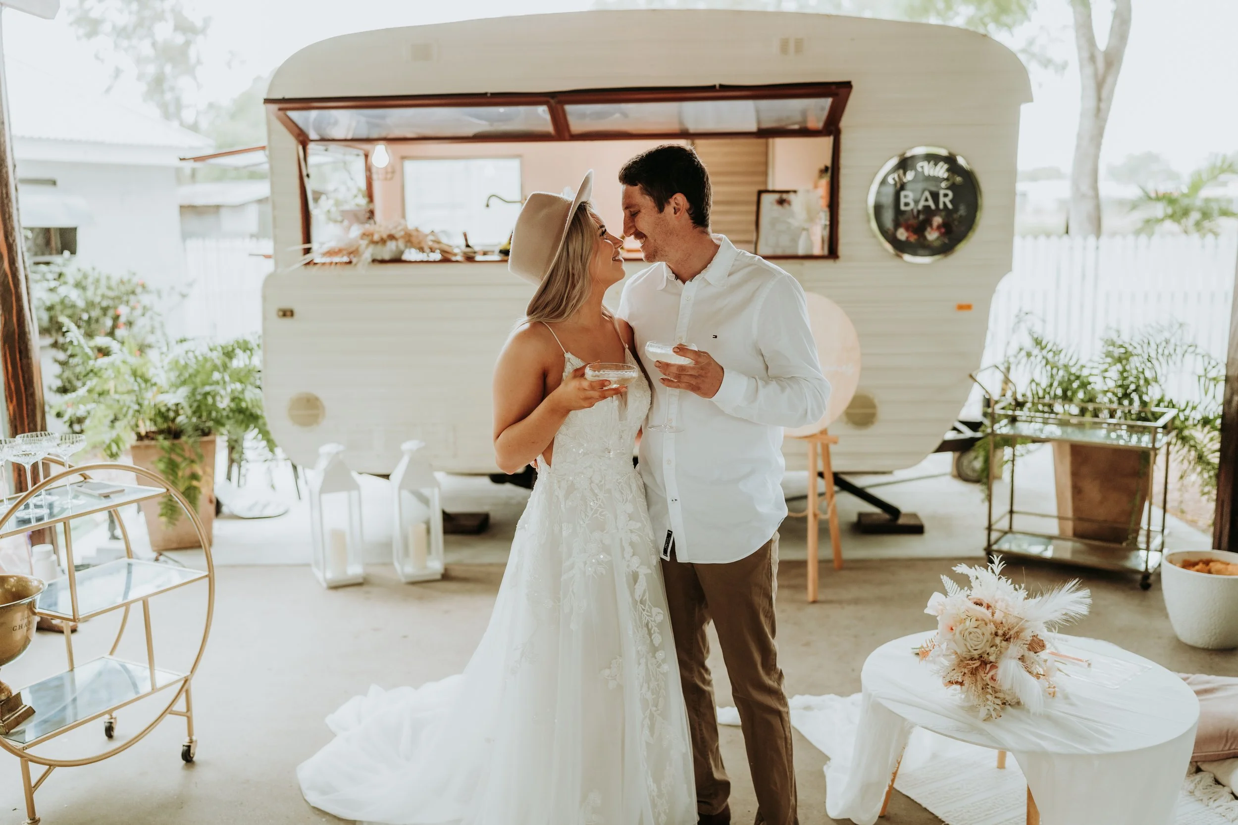 A bride and groom holding drinks and smiling at each other in front of a vintage trailer with a bar sign, decorated with flowers and greenery for a wedding celebration.