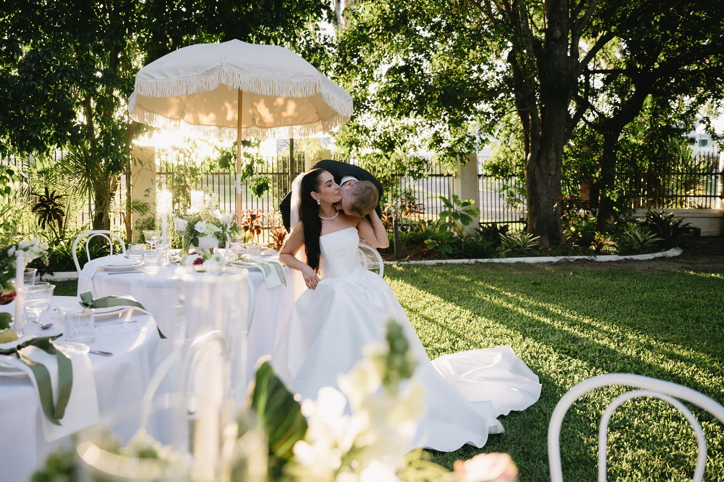 A bride in a white wedding dress and a veil sits at an outdoor wedding reception, kissing a man who is leaning over her. The reception area features a table with a white tablecloth, floral centerpieces, glasses, and tableware, shaded by a large white