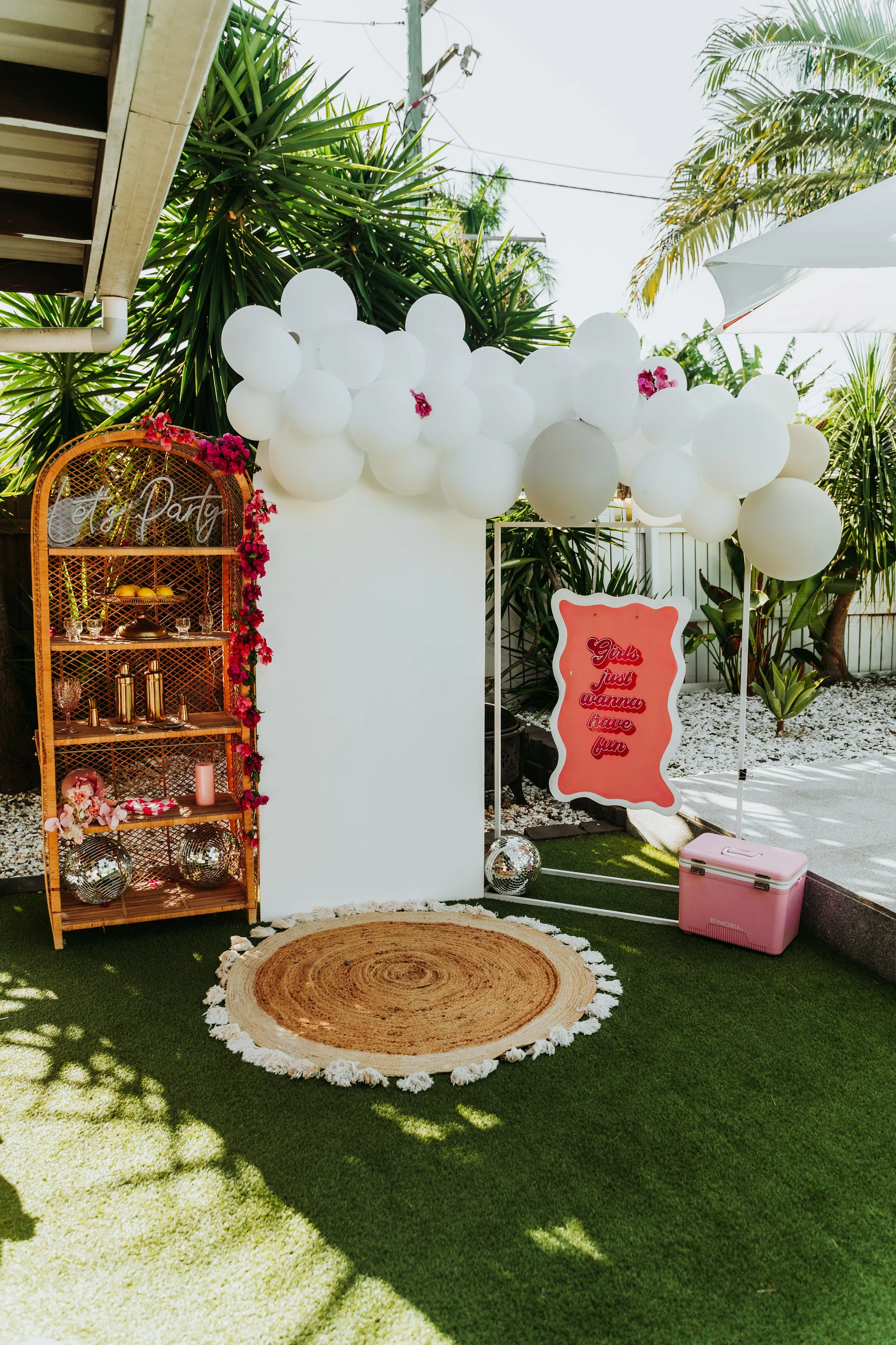 Decorative area with white balloons, pink flowers, a round woven rug, a pink sign with red lettering that reads 'Girls just wanna have fun,' a pink cooler, and a wooden shelf with gold and pink decor, set outdoors with lush green plants.