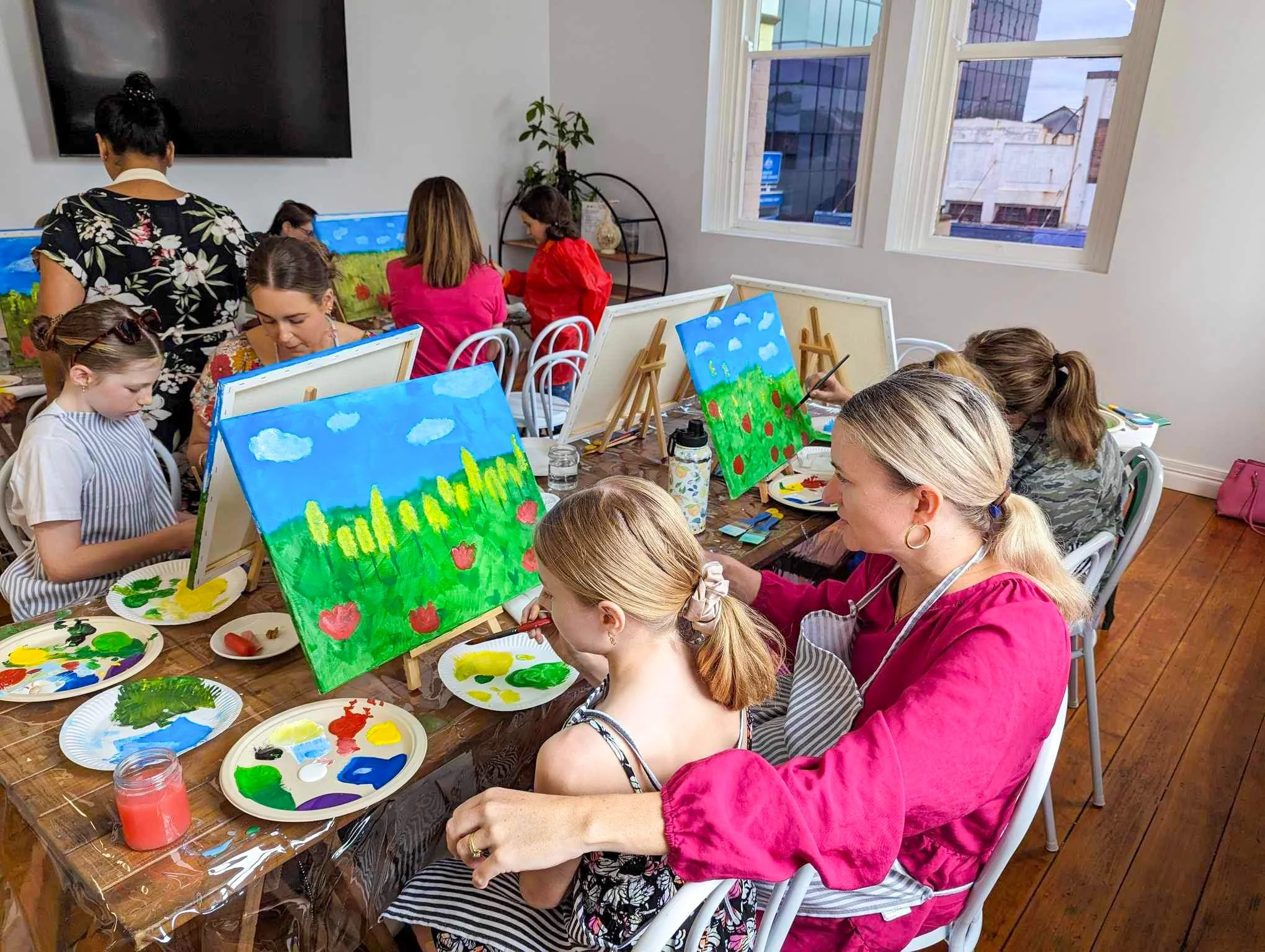 People, including children and adults, participating in a painting class, creating colorful landscape artworks on canvases set on easels, in a bright room with windows and wooden flooring.
