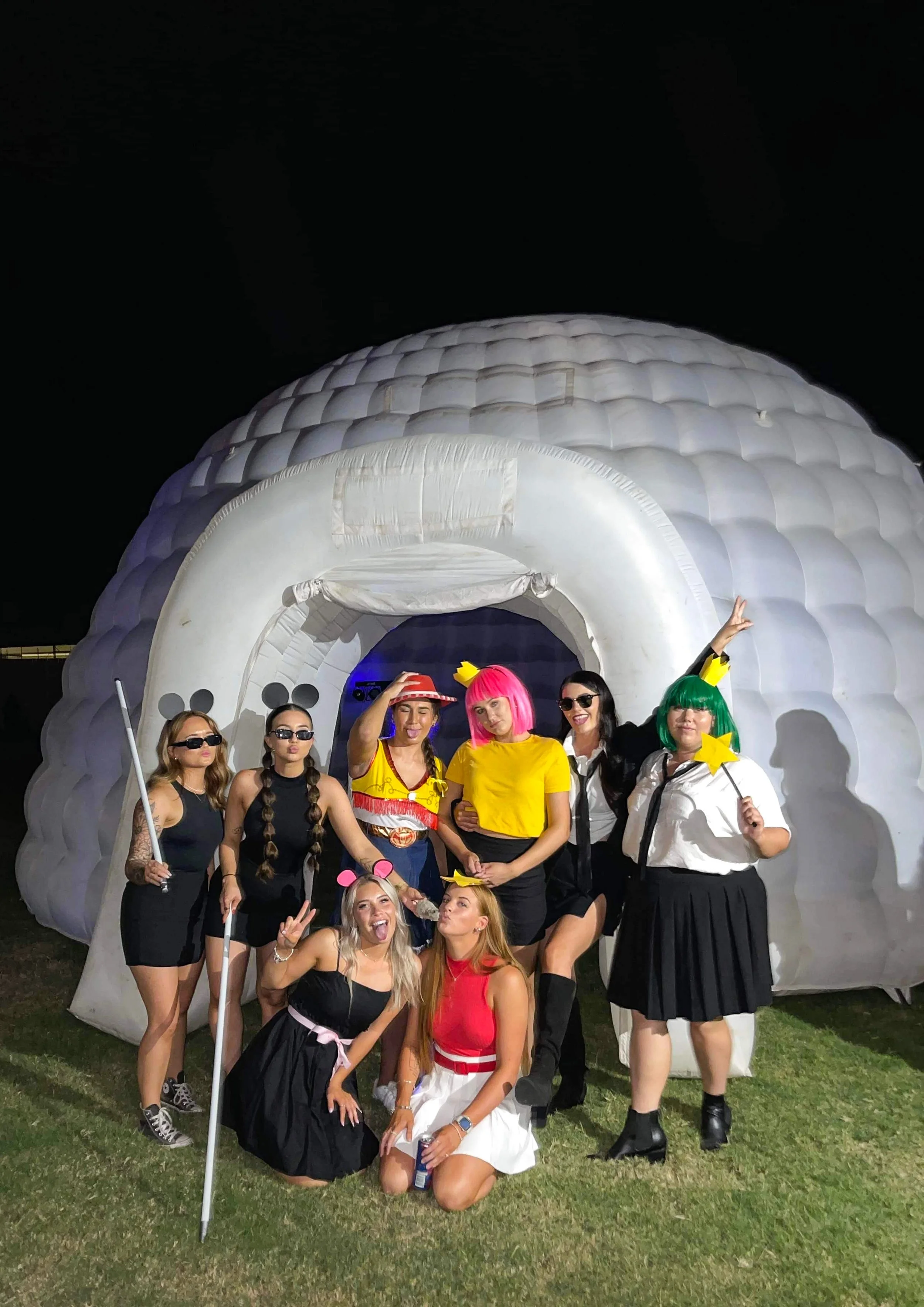 Group of women in colorful costumes posing in front of a large inflatable igloo at night.