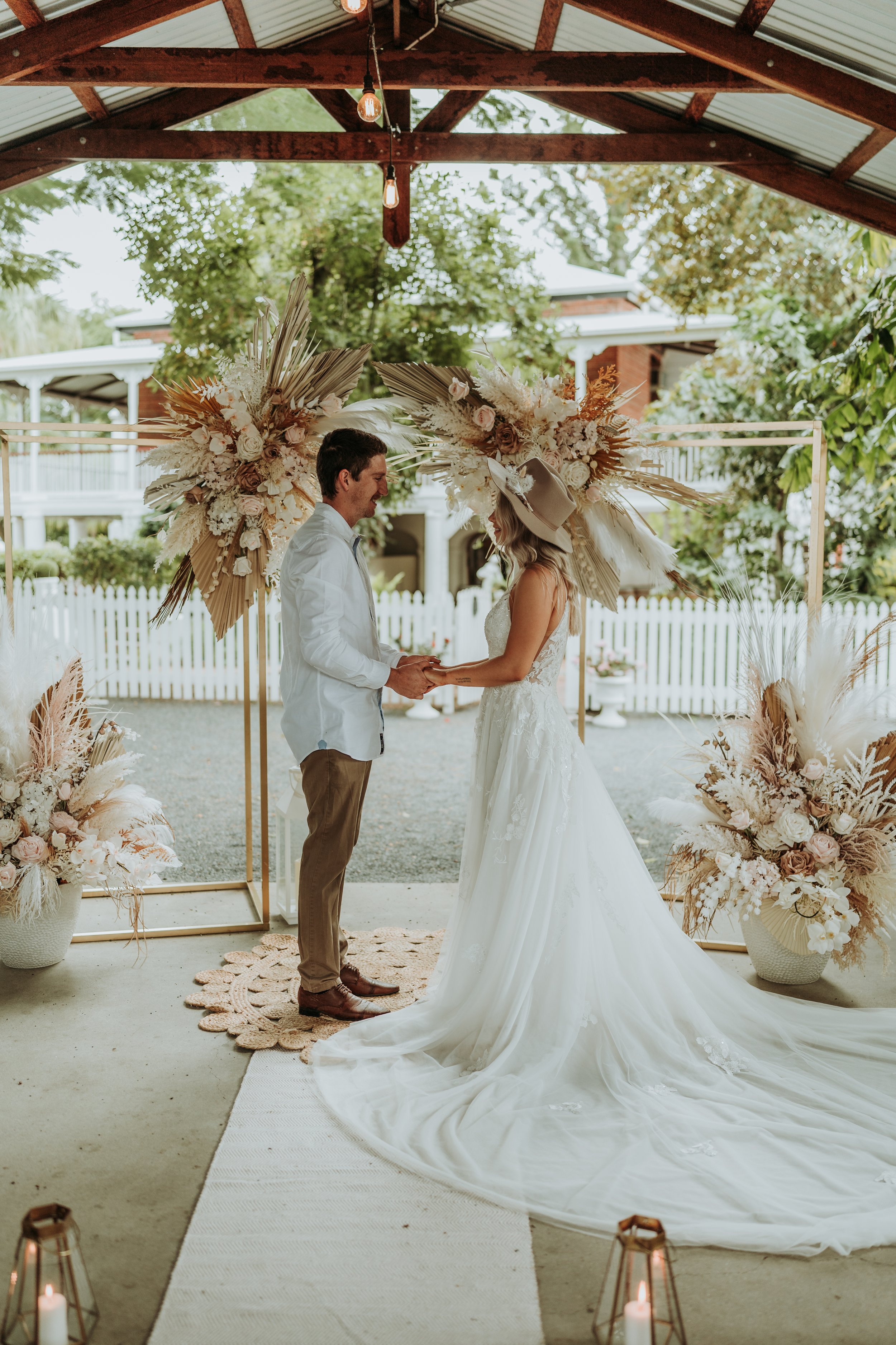 A couple exchanging vows during their wedding ceremony in an outdoor pavilion, decorated with large floral arrangements and candles.