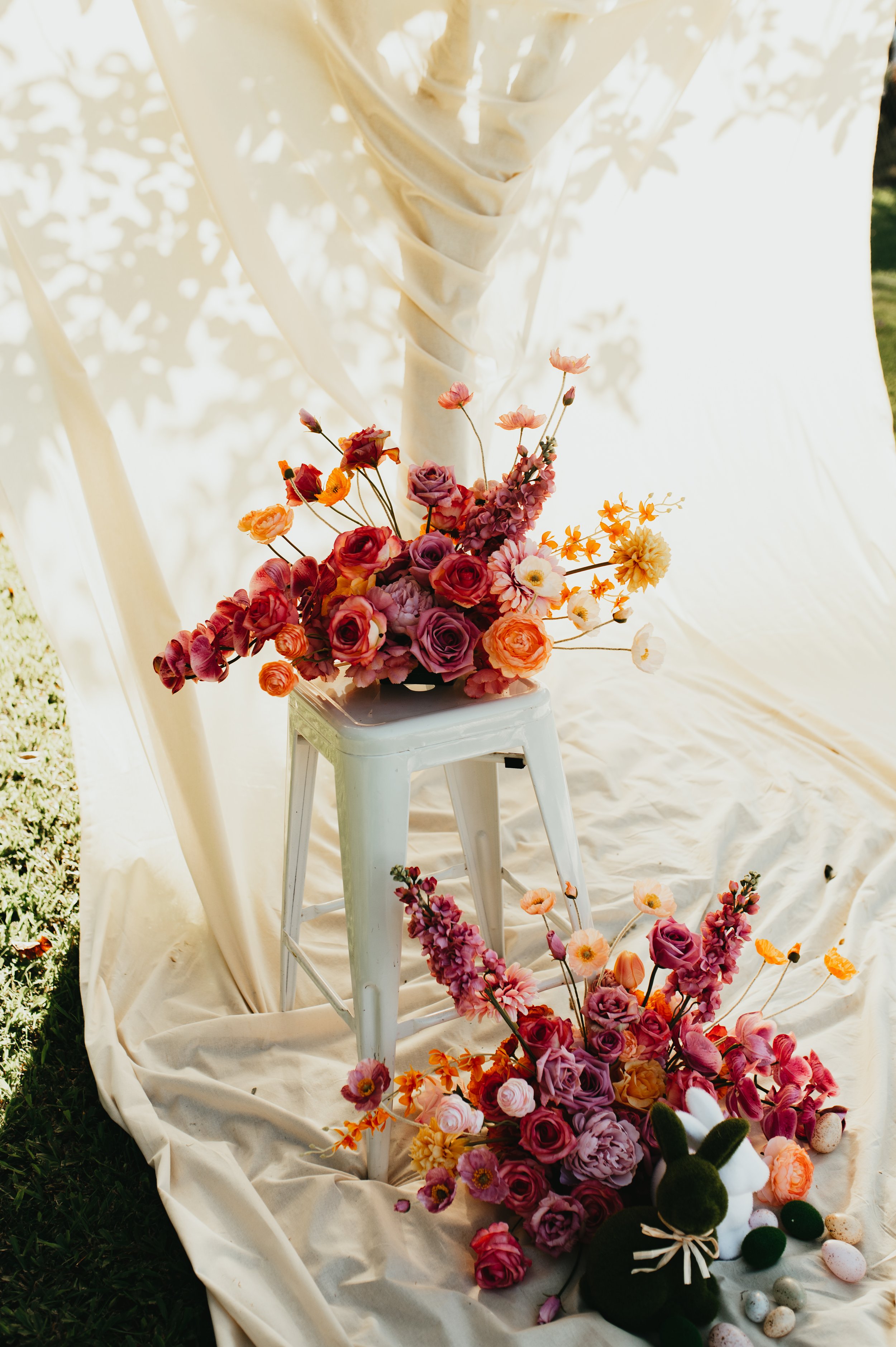 Arrangement of pink and purple flowers on a white stool and on the ground, with a cream-colored fabric backdrop, outdoor setting, and small decorative items nearby.