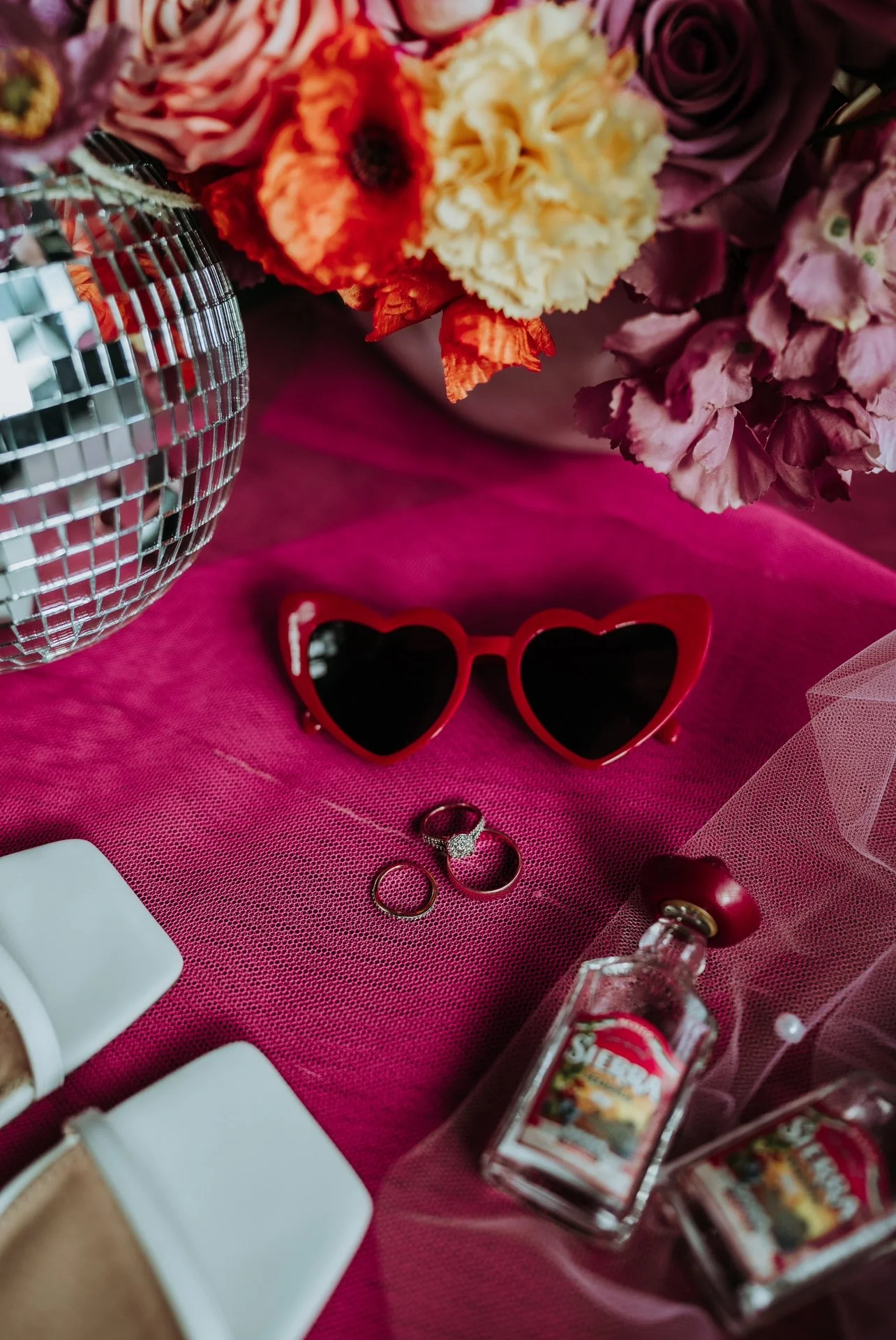 A pair of red heart-shaped sunglasses, a set of rings, a bottle of small perfume, white shoes, and a bouquet of flowers in a vase, all arranged on a pink surface.