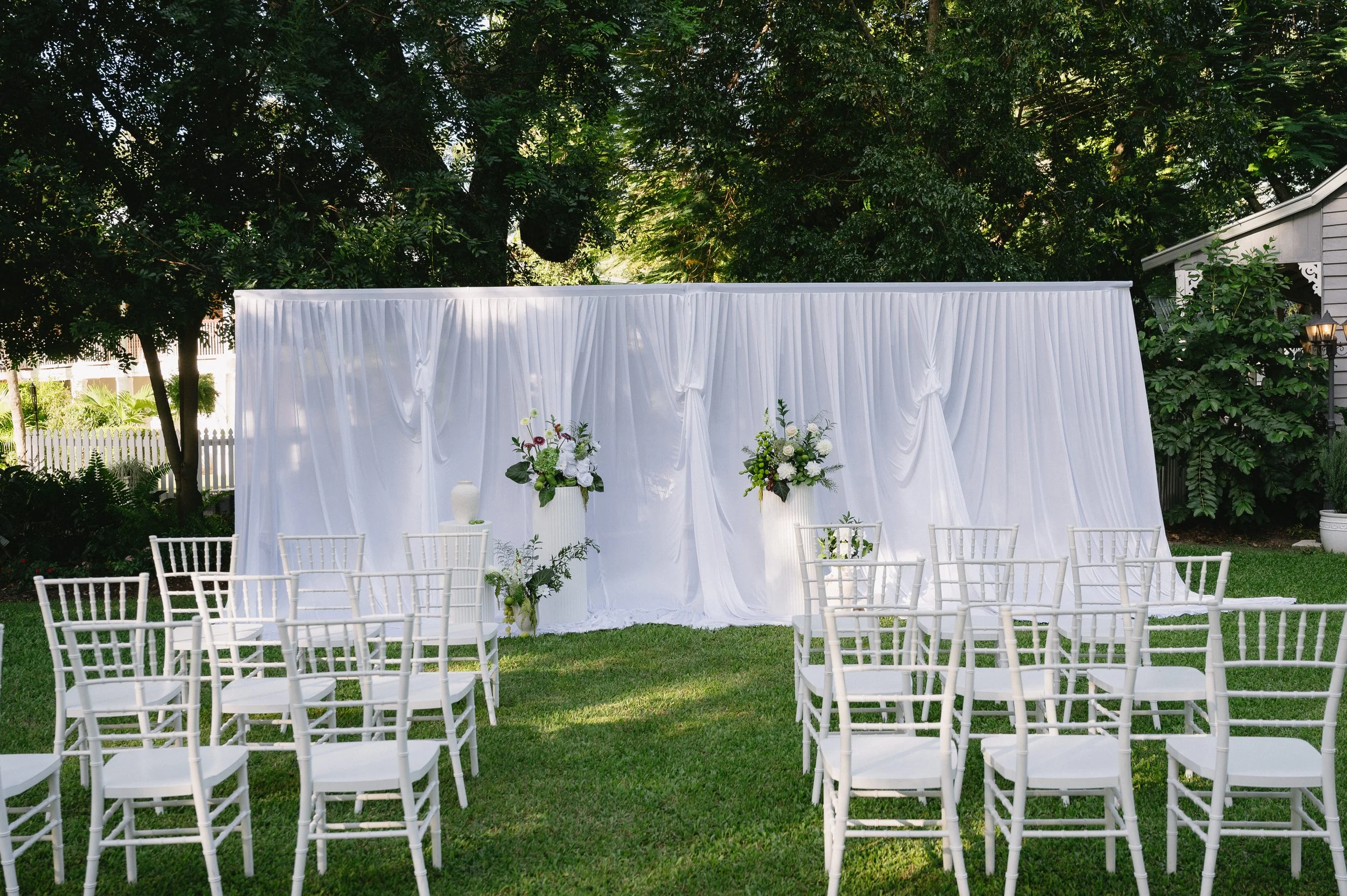 Outdoor wedding or event setup with white chairs arranged on grass facing a white draped backdrop decorated with floral arrangements, set in a garden with trees and a house nearby.