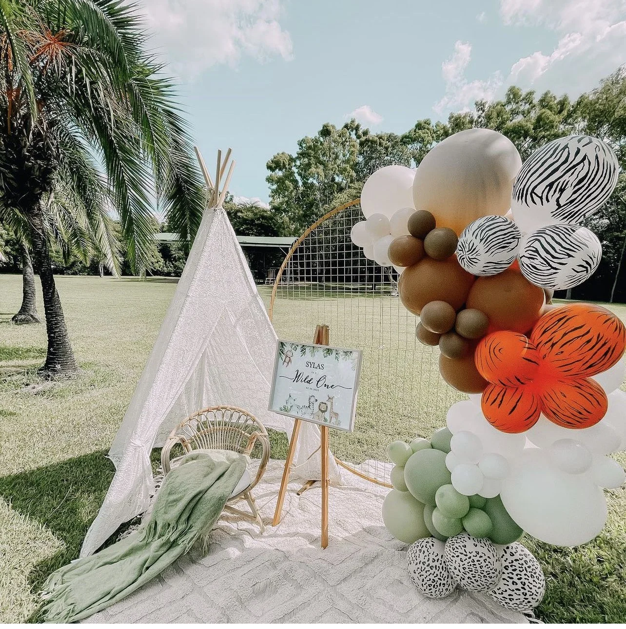 Decorative outdoor setup with a white teepee, a rattan chair with a green blanket, a sign on an easel reading 'Sylas Wild One,' and a large balloon arrangement with animal print and solid-colored balloons, set on a grassy field with trees and a clear sky.