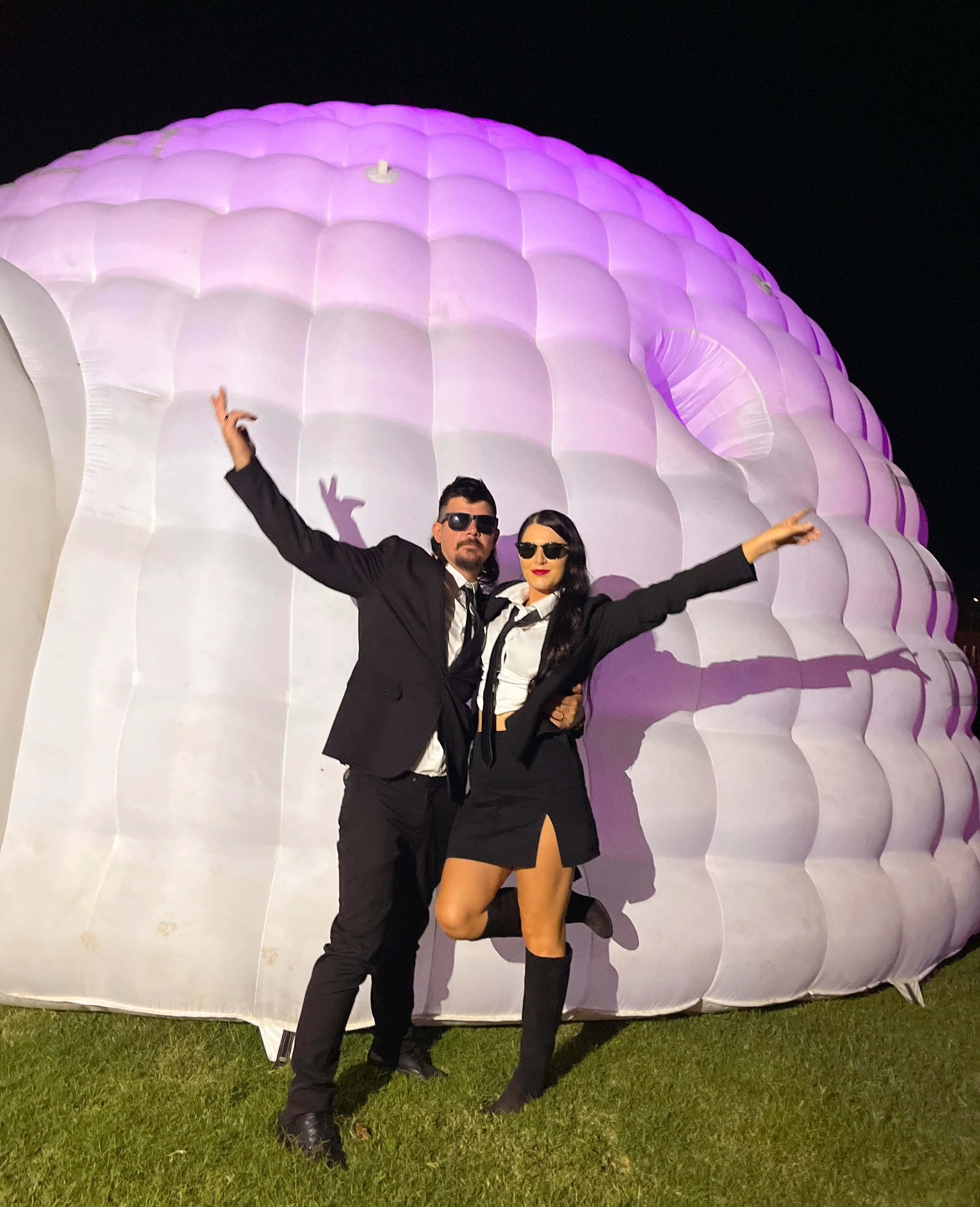 Two individuals dressed in black suits and sunglasses are posing in front of a large, illuminated, purple and white inflatable structure at night. The woman is wearing a black skirt, white shirt, black tie, and knee-high boots, while the man is weari