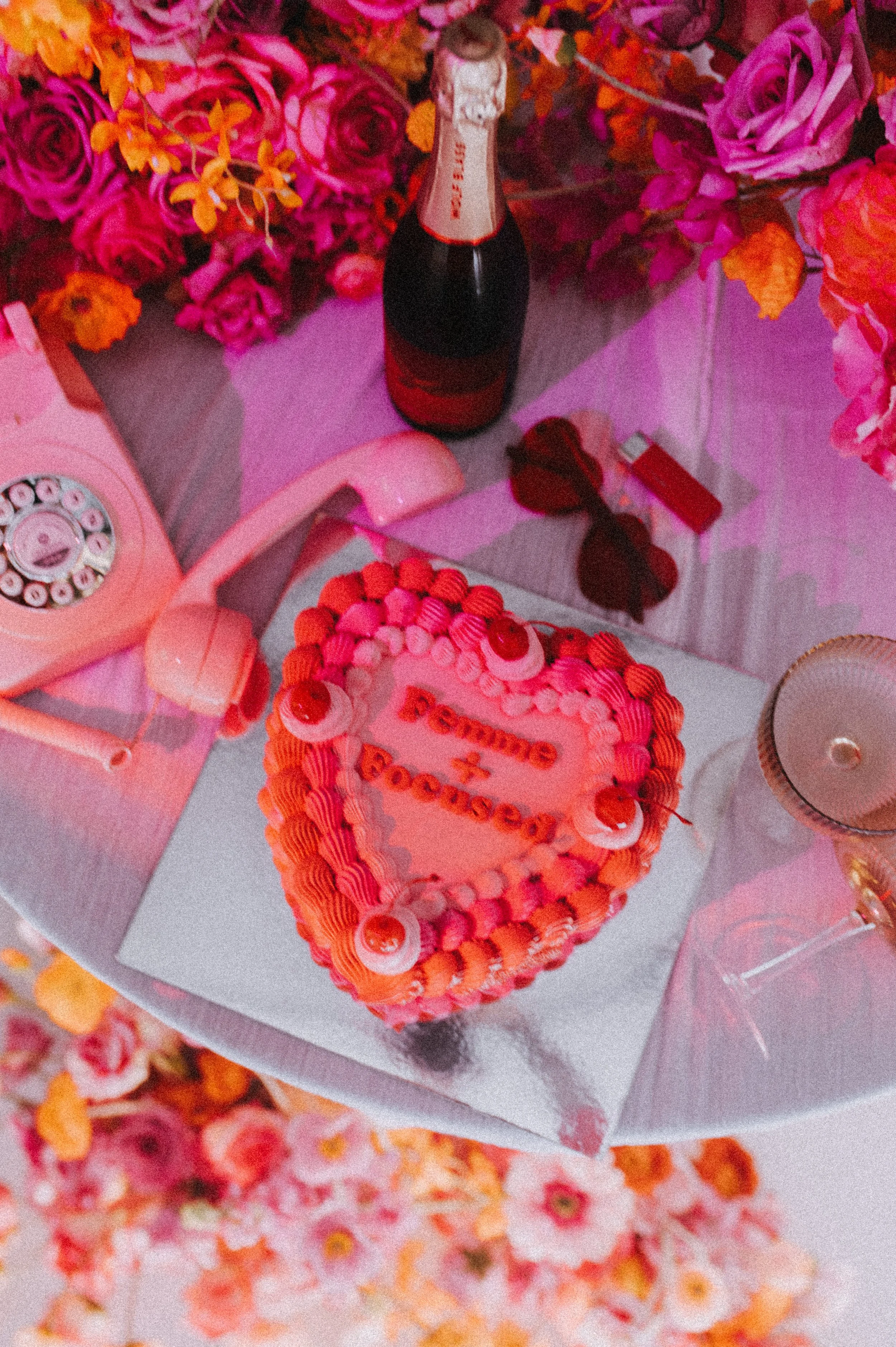 Heart-shaped cake with orange and pink frosting, decorated with cherries and the words "Some & Forever" written on top, surrounded by a pink floral arrangement, a pink rotary phone, a bottle of champagne, sunglasses, a lighter, and a glass on a table.