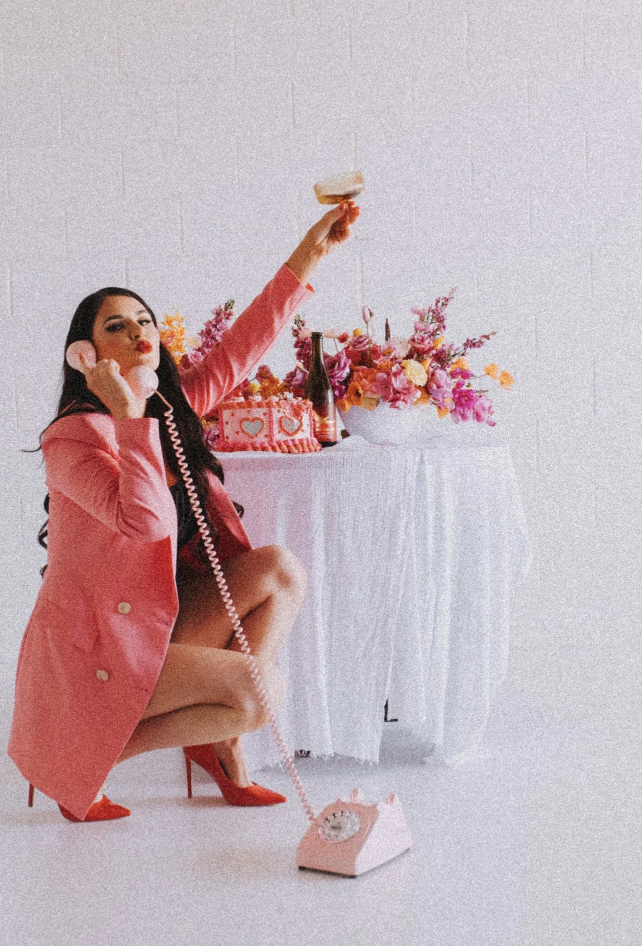 Woman in pink blazer and red high heels squatting while holding a pink rotary phone, with one arm raised holding a doughnut, in front of a table with a pink birthday cake, flowers, and drinks.