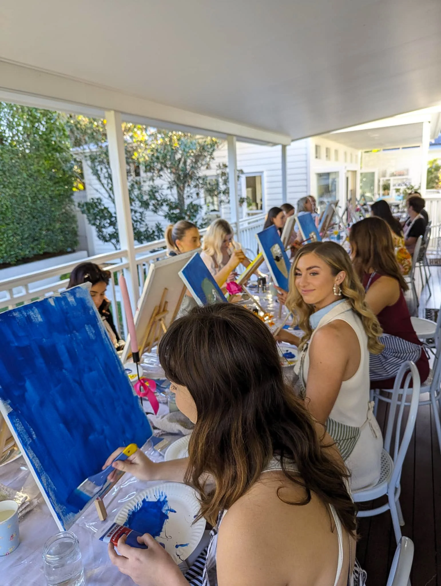 Women participating in an outdoor painting class on a porch, with canvases and paint supplies in front of them.