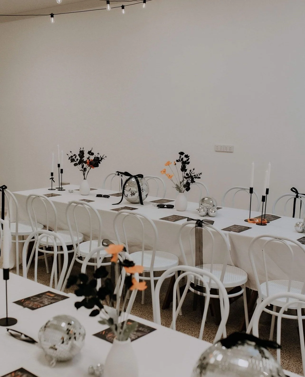Empty banquet table decorated with vases of dark and light flowers, candlesticks, disco balls, and place settings, set for a celebration.