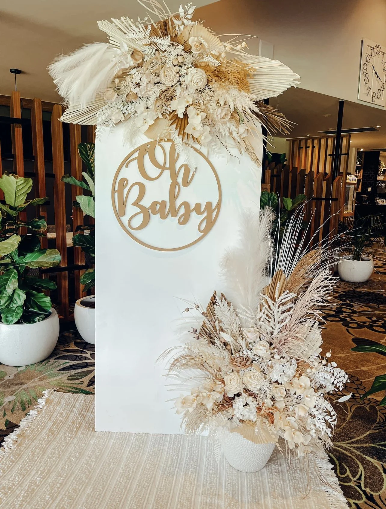 A decorative white panel with a sign that says 'Oh Baby' in cursive, adorned with cream, beige, and gold dried floral arrangements at the top and bottom. The setup is in an indoor space with potted plants and a patterned carpet.