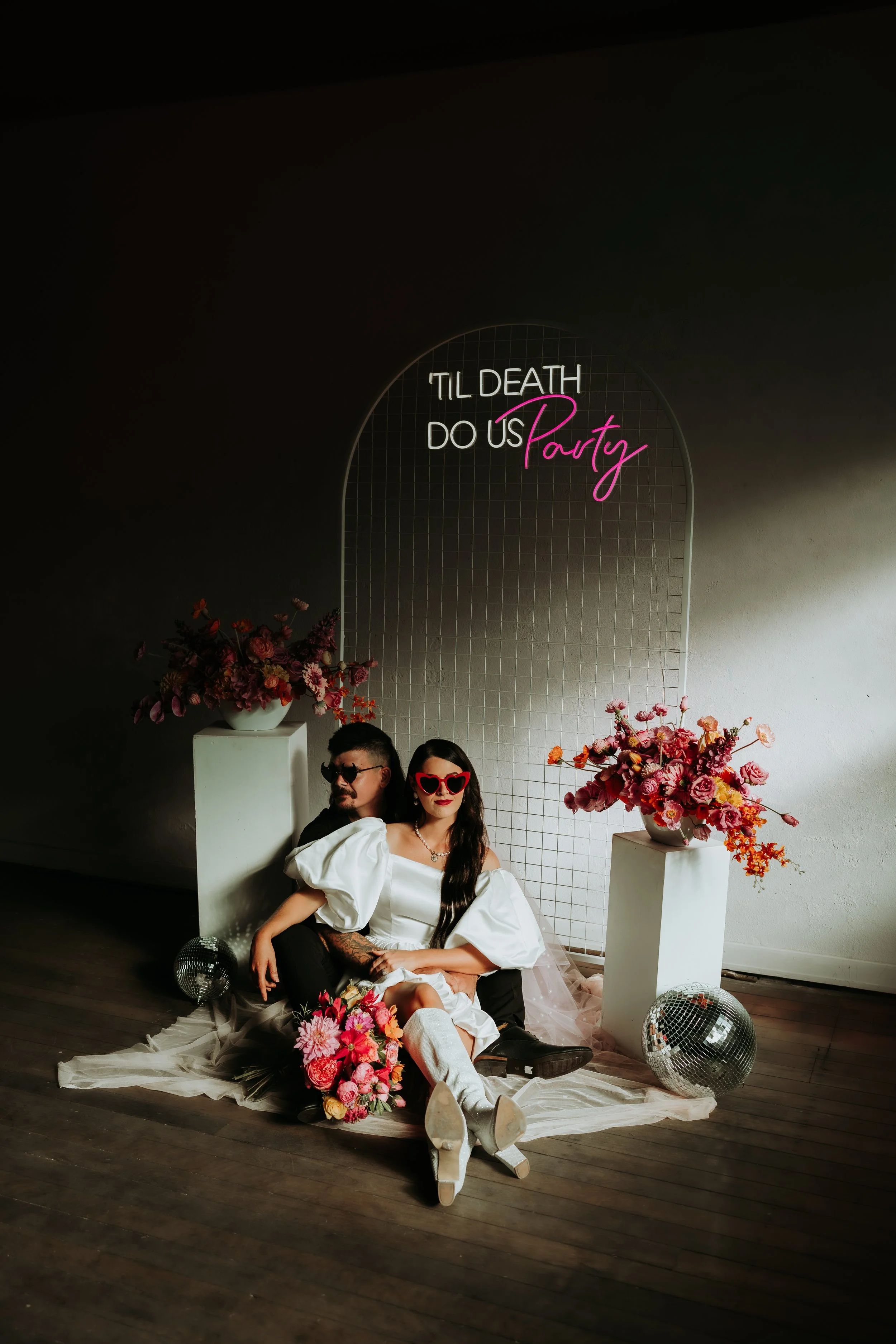 A couple sits on the floor in front of a mirror with neon sign that says '’Til Death Do Us Party', surrounded by white pedestals with pink and purple flowers, disco balls, and a white fabric backdrop.