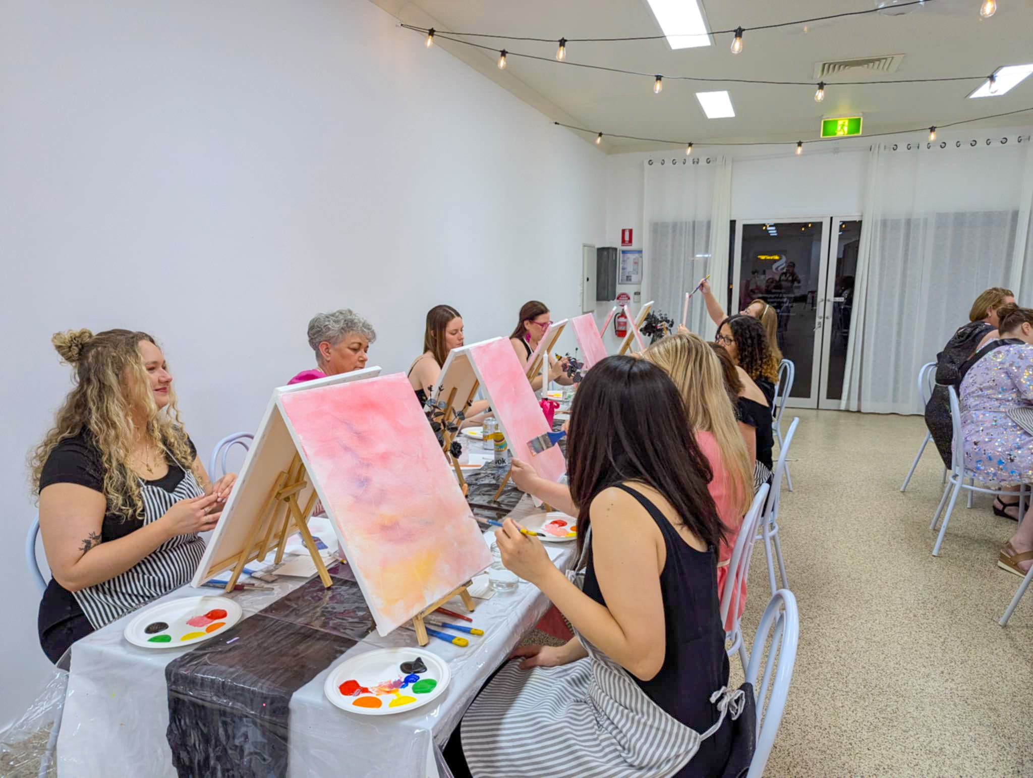 A group of women participating in a painting class, sitting at a long table with art supplies, creating colorful abstract paintings on canvases.