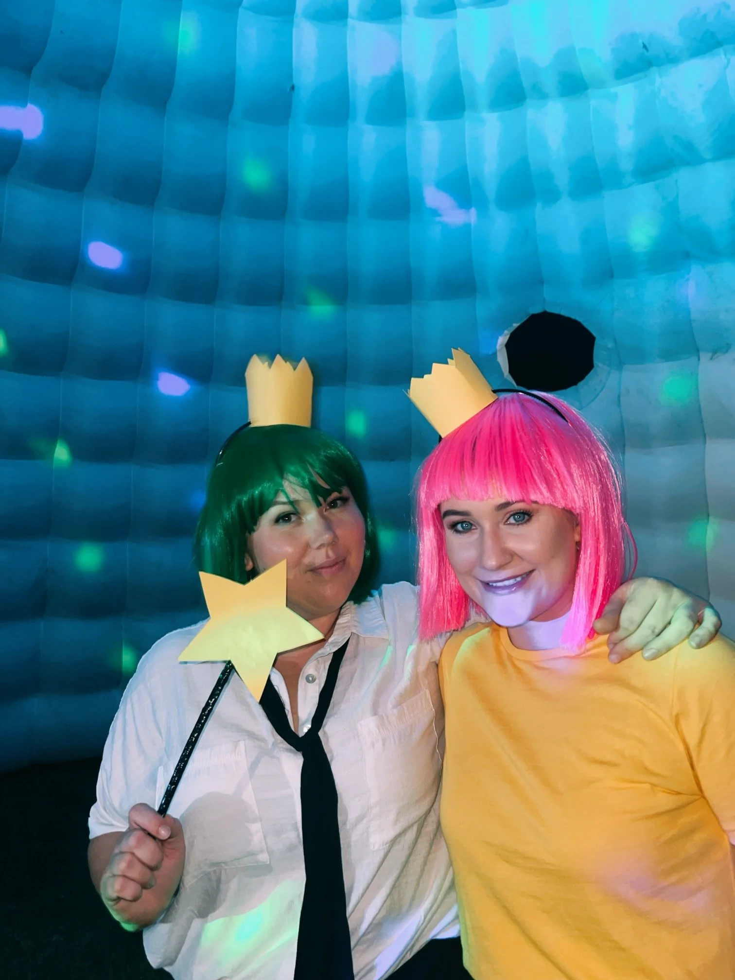 Two women with colorful wigs and paper crowns, one with a green wig and the other with a pink wig, posing together with a blue, illuminated, textured background.